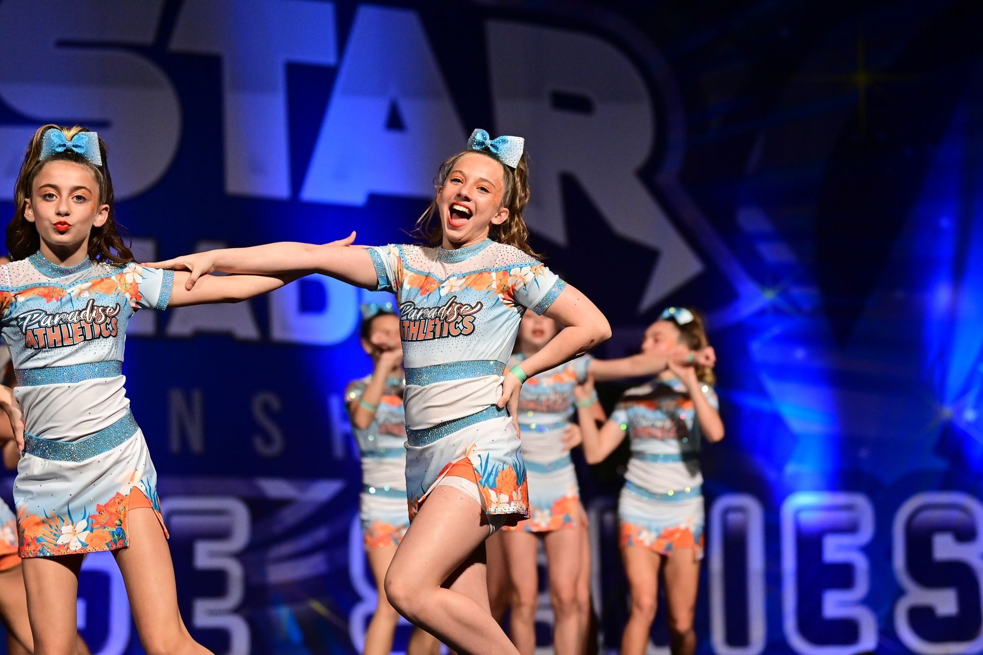 A group of cheerleaders in light blue and orange uniforms perform on stage during a competition.