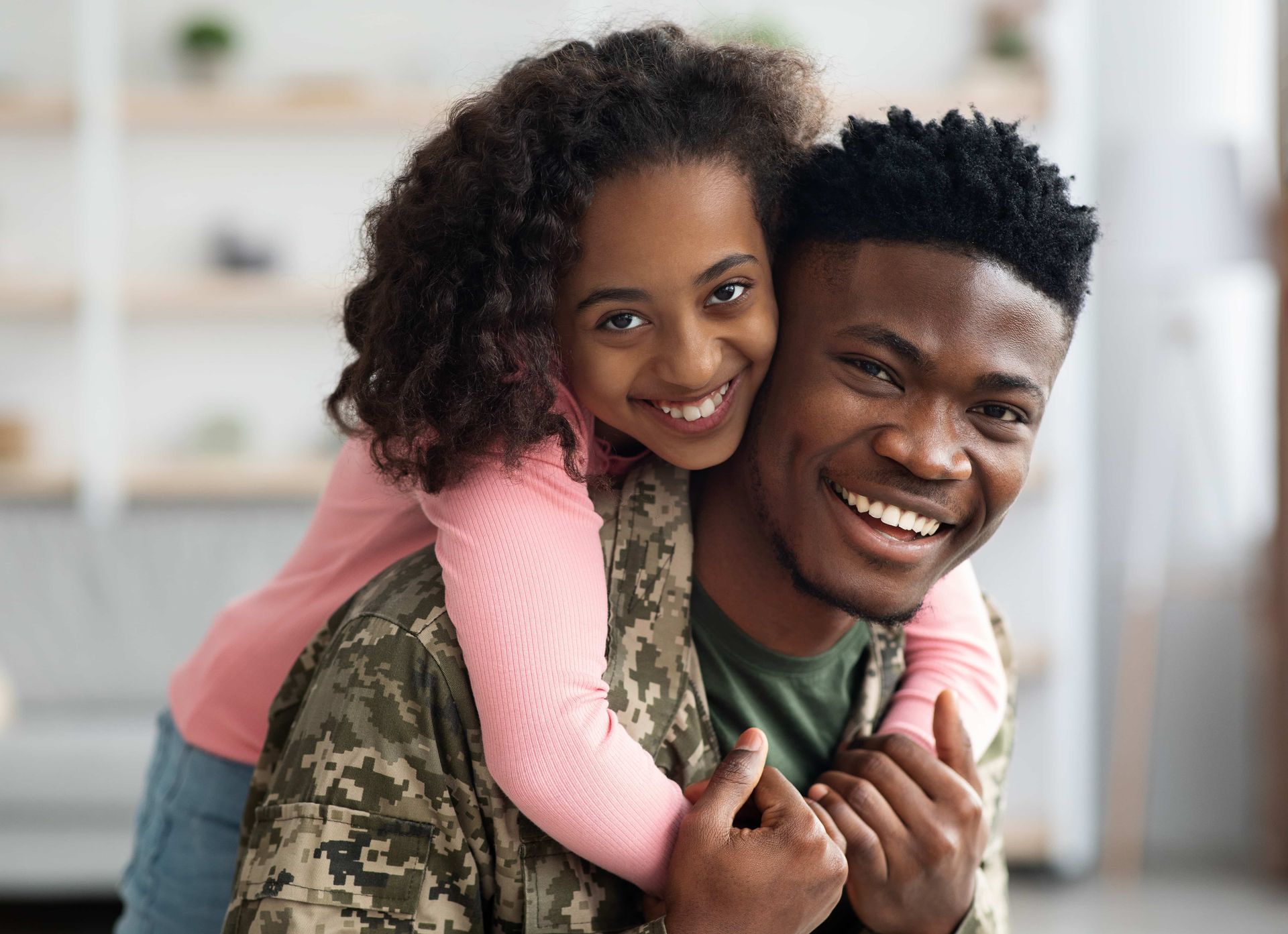 Smiling couple indoors, the woman hugging the man from behind on a couch.