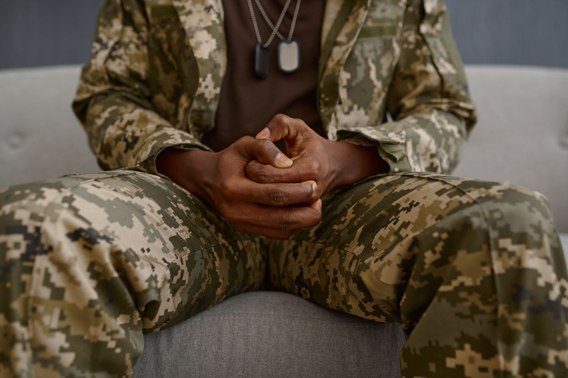 Person in camouflage uniform seated on a couch with hands clasped, wearing dog tags.
