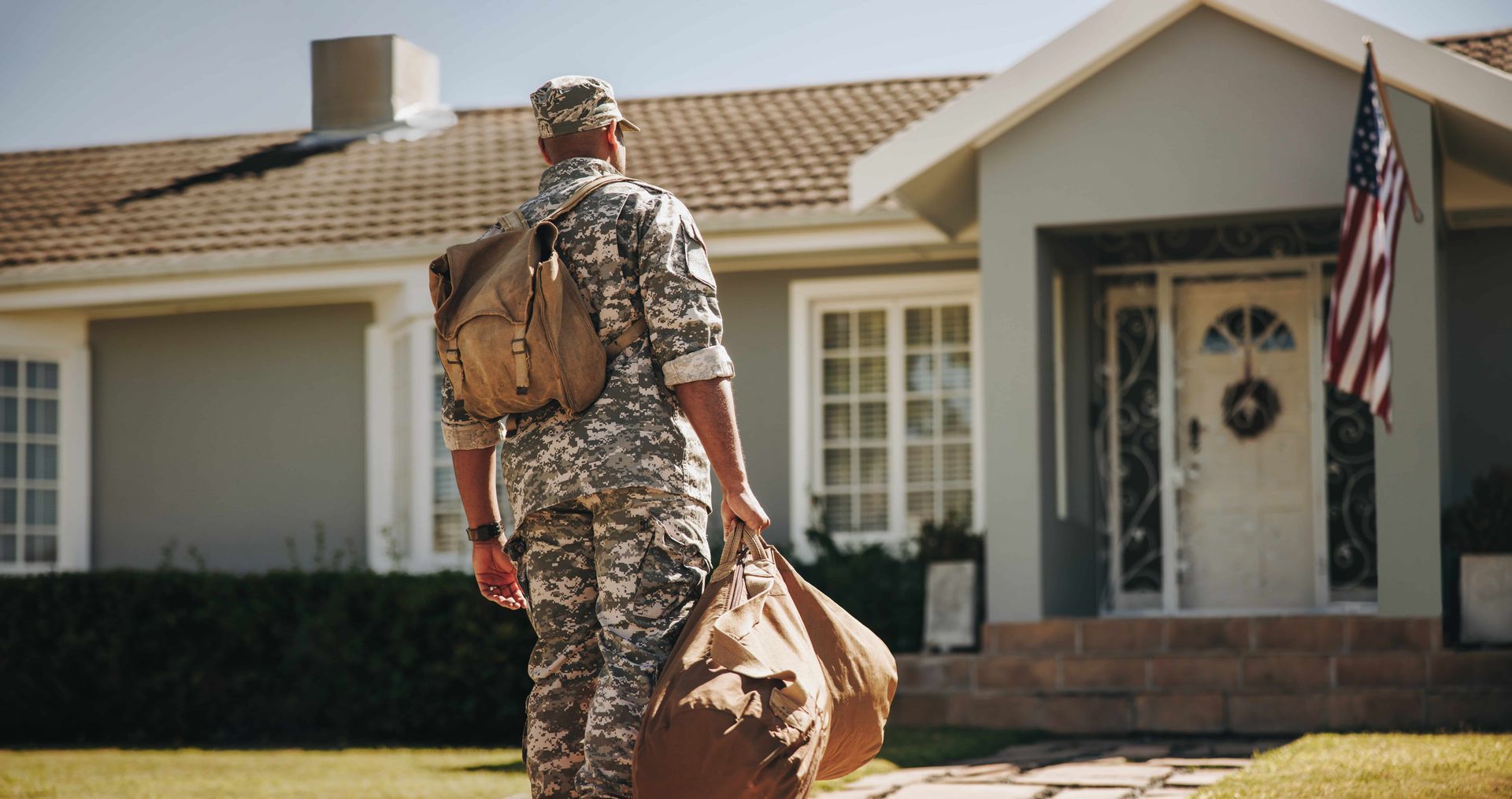 Soldier in camouflage carrying a duffel bag walks toward a suburban house with an American flag.