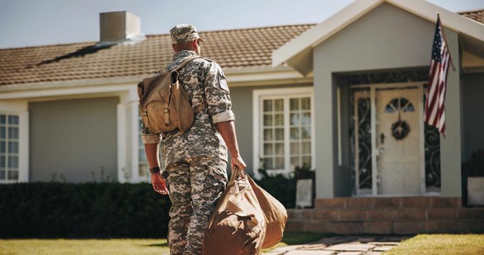 Soldier in camouflage carrying a duffel bag walks toward a suburban house with an American flag.