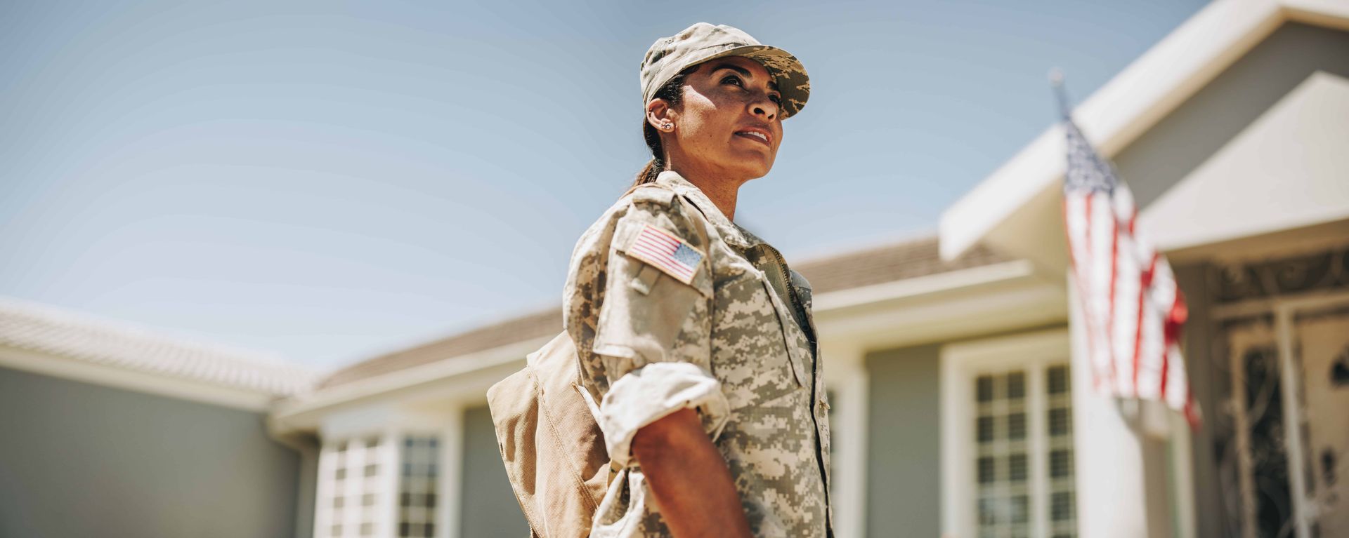 Person in military uniform standing outside a house near an American flag under a sunny sky