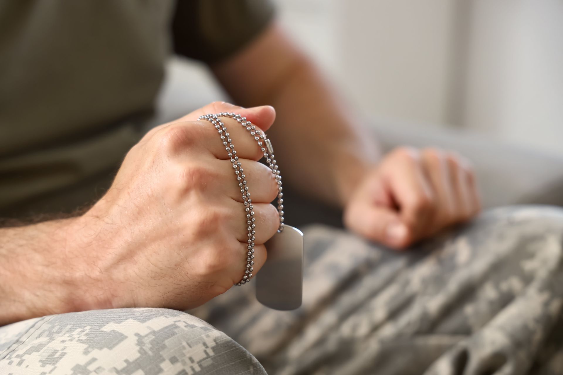 Person holding dog tags in a camouflage setting, with another person blurred in the background