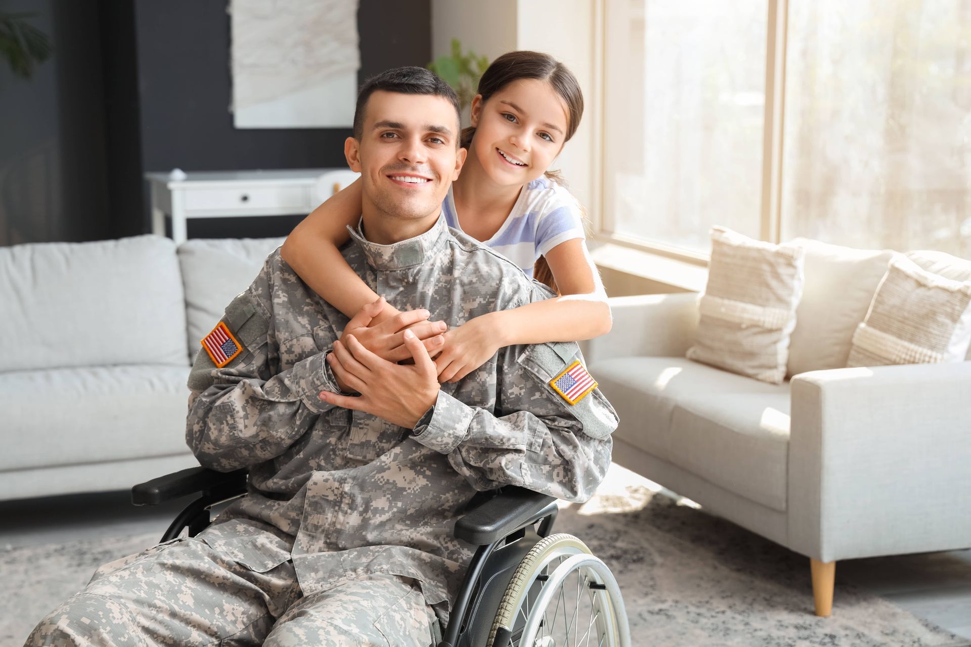 Man in a wheelchair smiling as a child hugs him from behind in a bright living room.