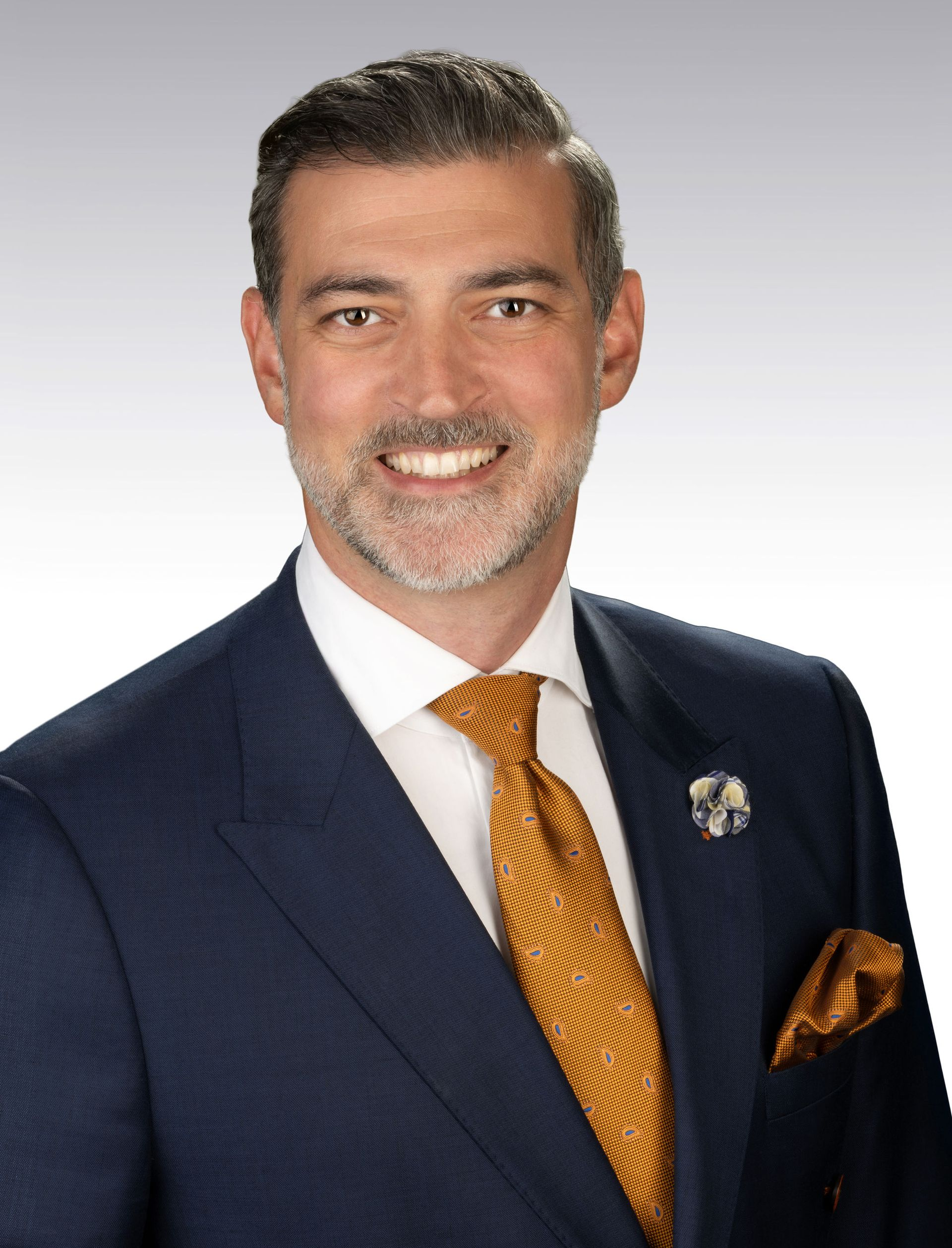 Man in navy suit, gold tie, smiling, pocket square, and lapel pin.