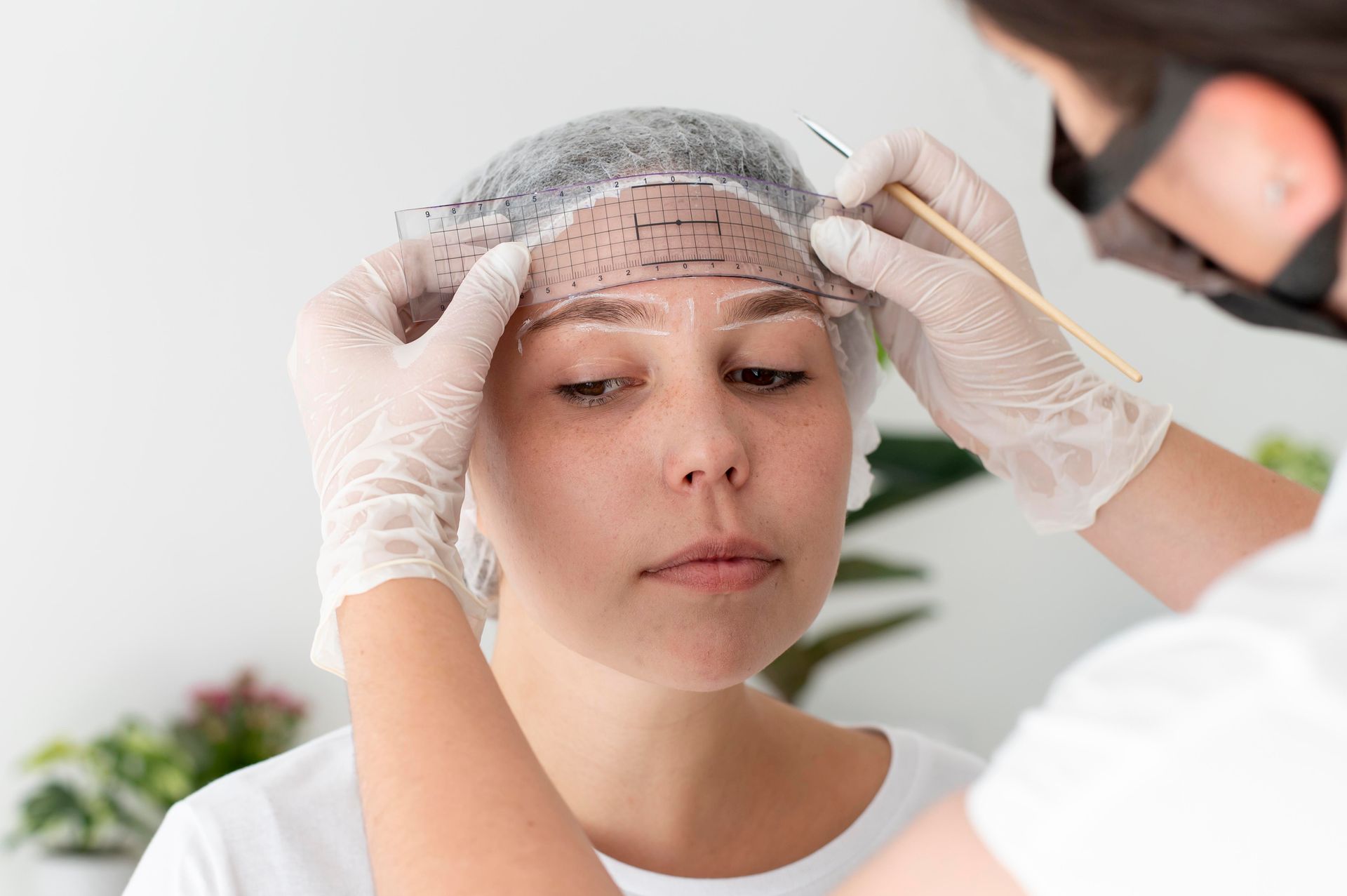 Person having eyebrows measured for shaping by a gloved technician. White background.