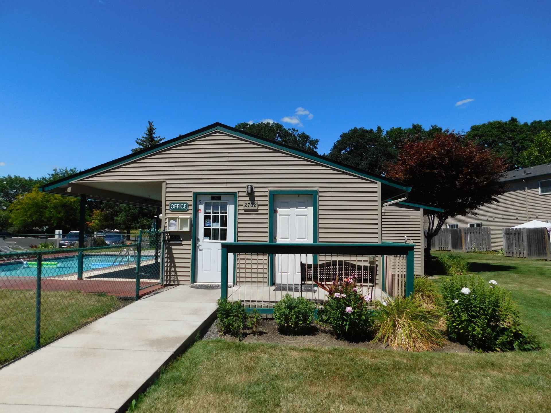 Poolside building with beige siding, green trim, and a white door, with a pool in the background.