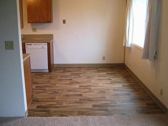Kitchen area with wood-look flooring, white dishwasher, brown cabinets, and a window with cream curtains.