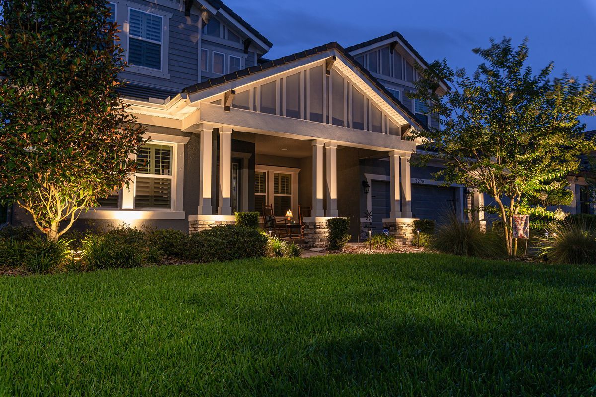 A warmly lit suburban house with a front porch at dusk, surrounded by a green lawn and trees