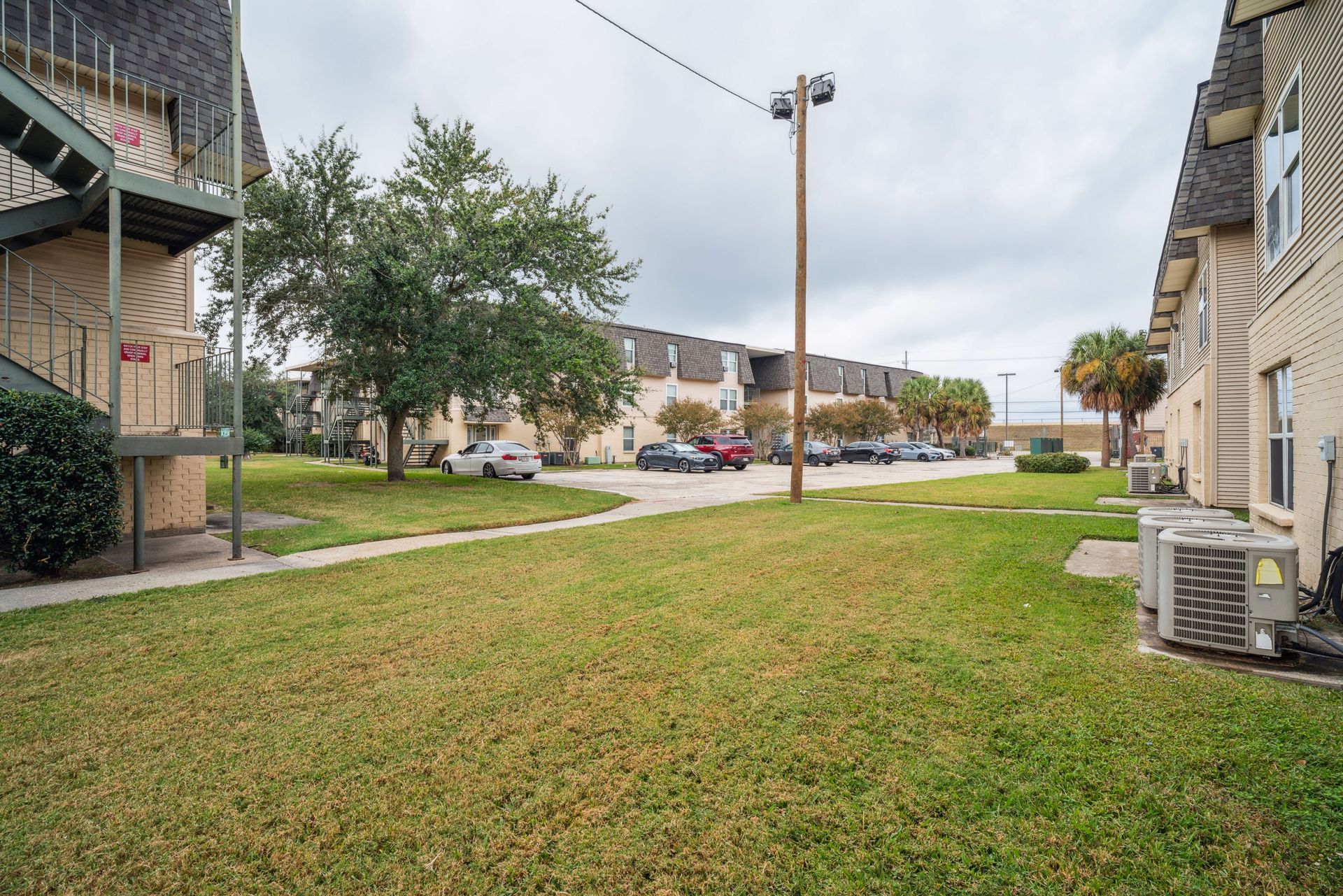 Apartment complex courtyard with green lawn, sidewalk, trees, and parked cars under a cloudy sky