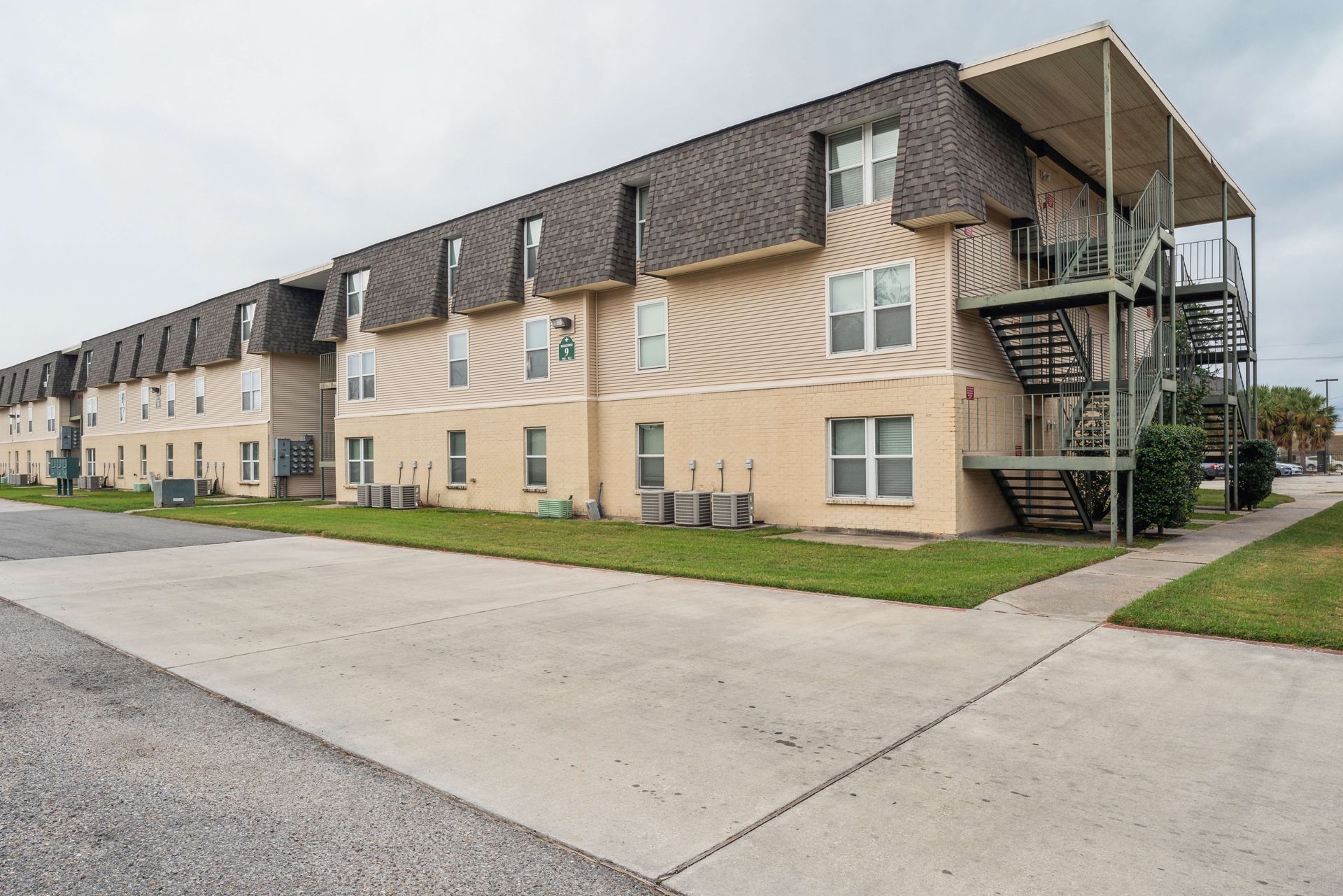 Beige apartment building with dark roof and exterior stairs beside a paved road.