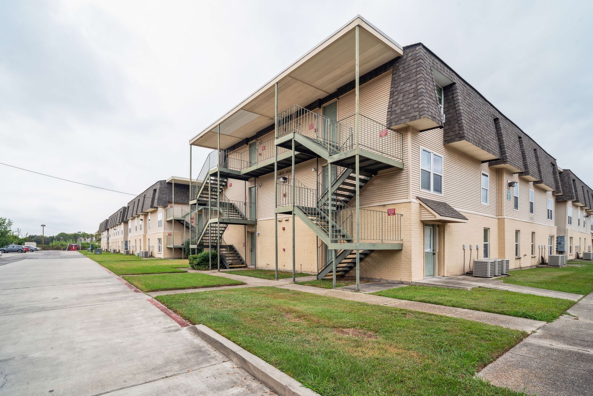 Apartment complex with beige siding, exterior stairs, and a sidewalk along a grassy parking lot