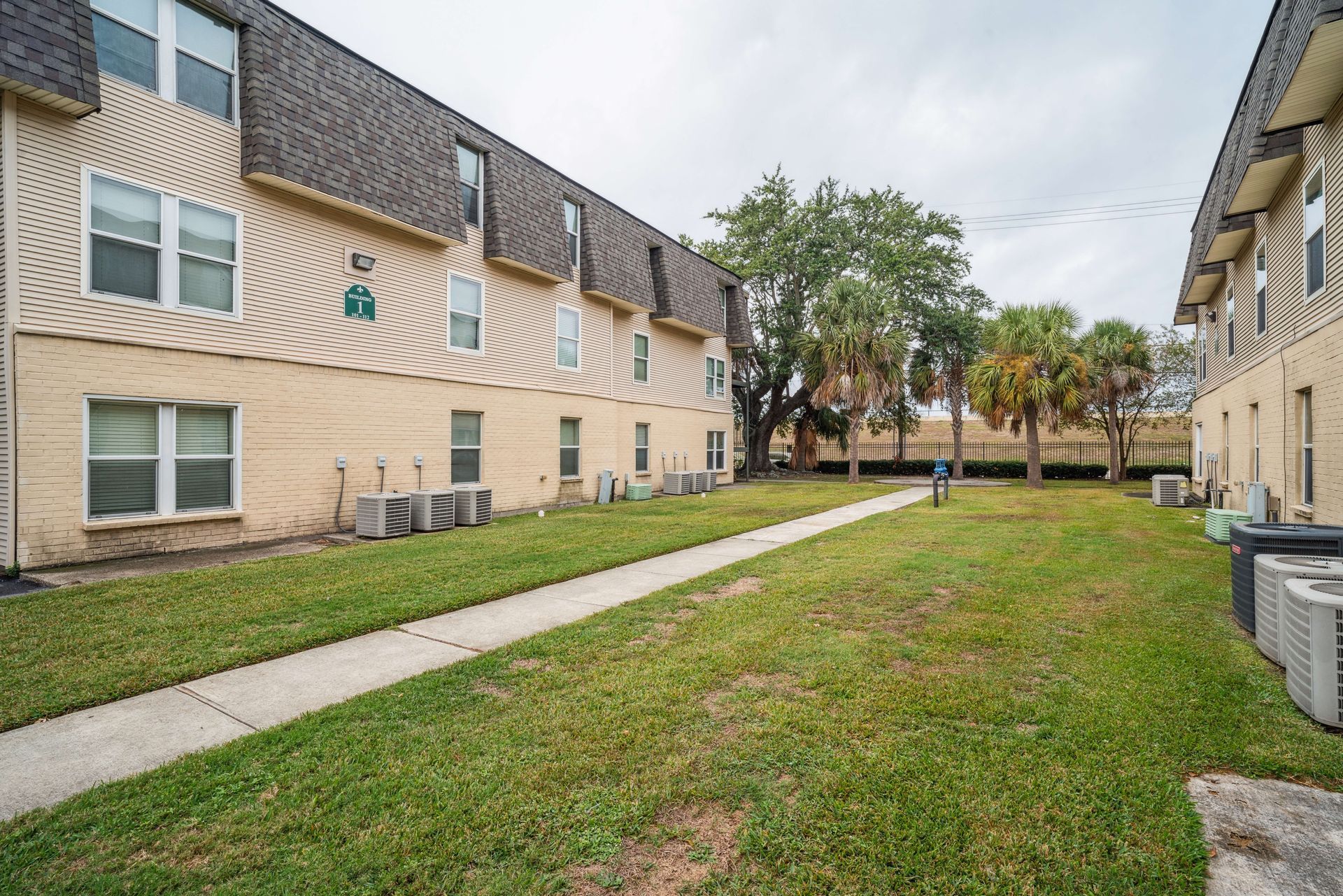 Apartment courtyard with beige buildings, grassy lawn, and sidewalk under a cloudy sky