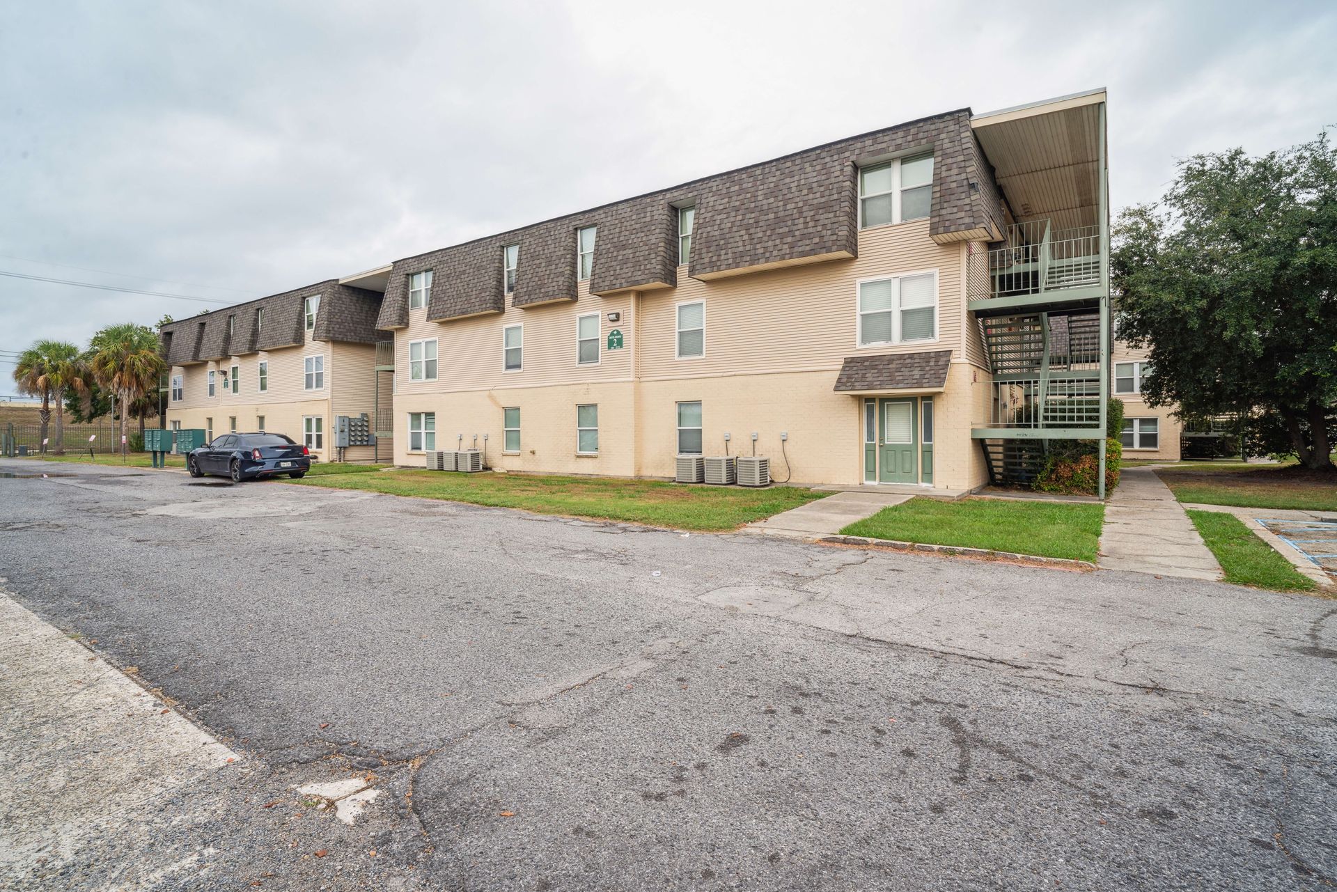 Apartment complex with beige siding, parked car, and gravel lot under cloudy sky