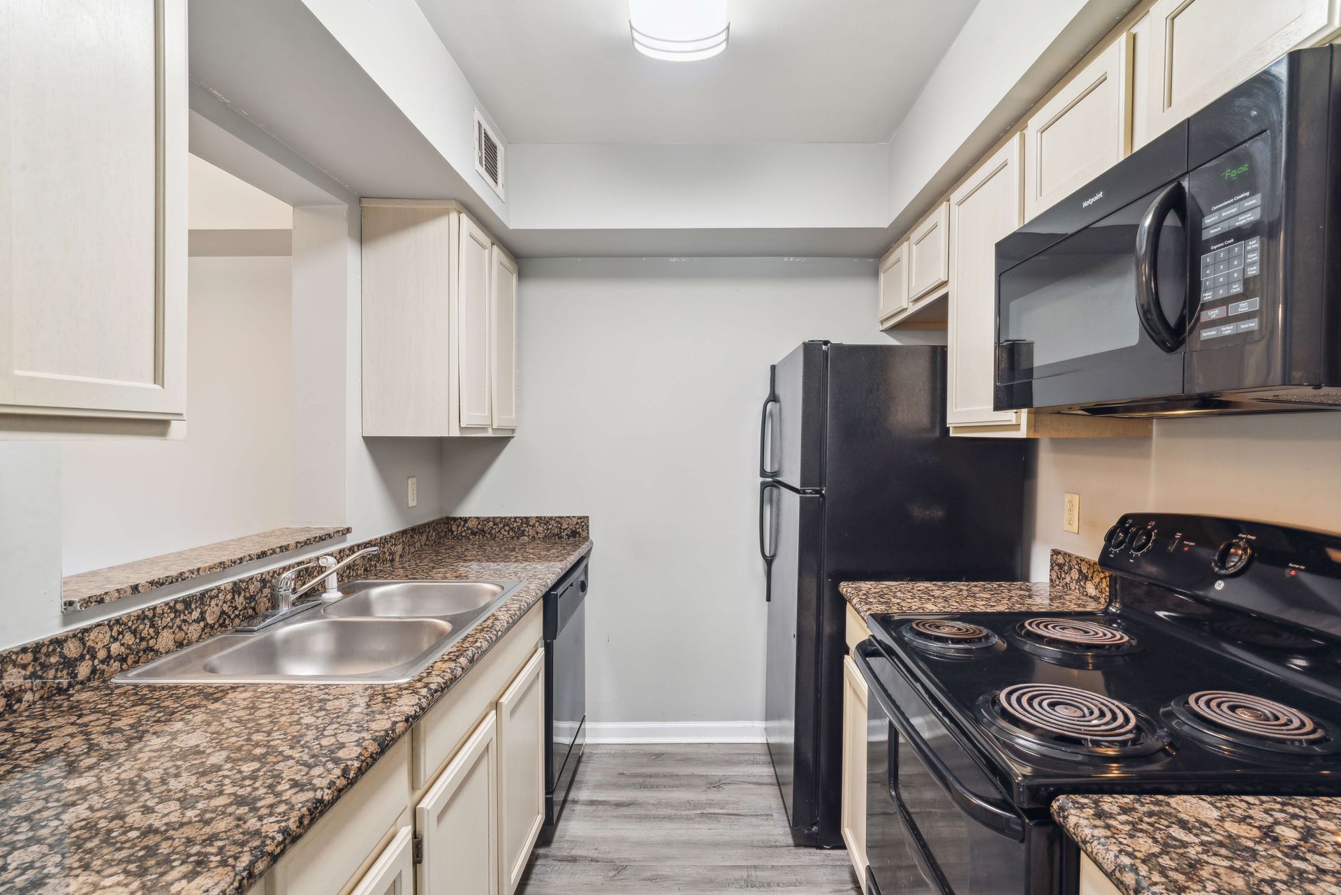Compact galley kitchen with beige cabinets, speckled countertops, black appliances, and white backsplash.