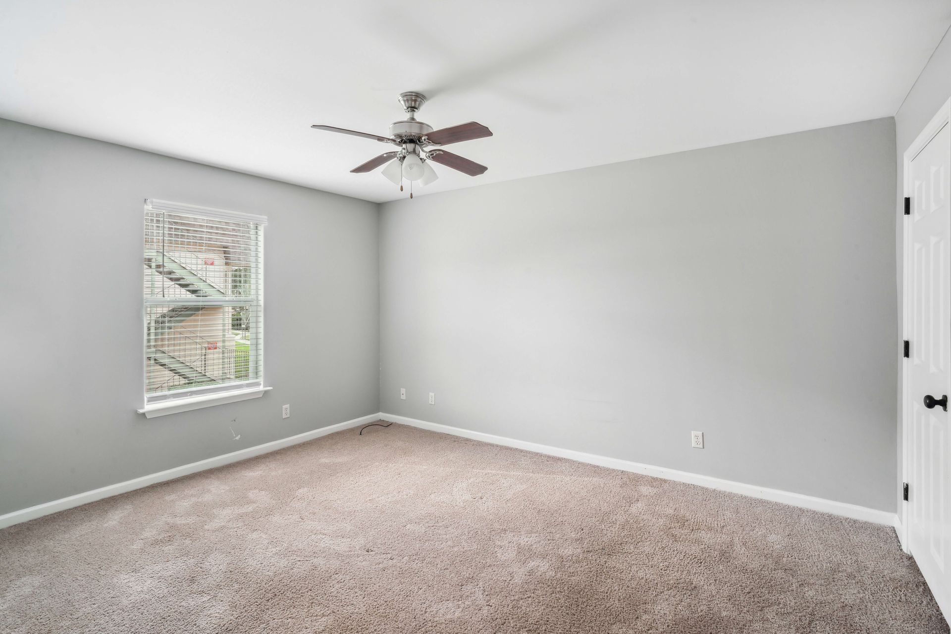 Empty gray carpeted bedroom with vaulted ceiling, ceiling fan, and window with blinds.