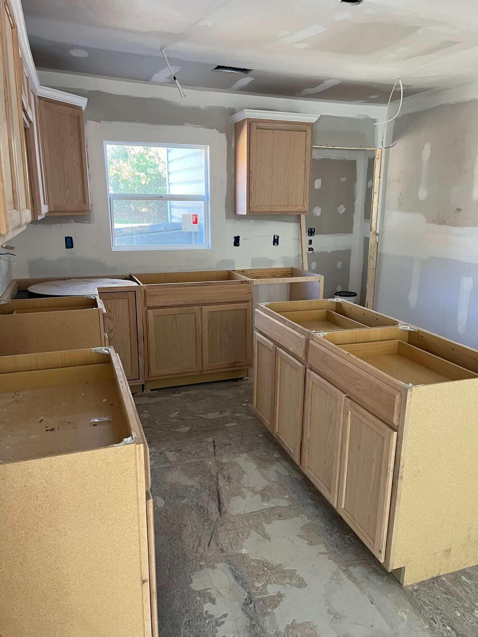 Kitchen cabinets in a room under construction; drywall, window, and unfinished flooring visible.