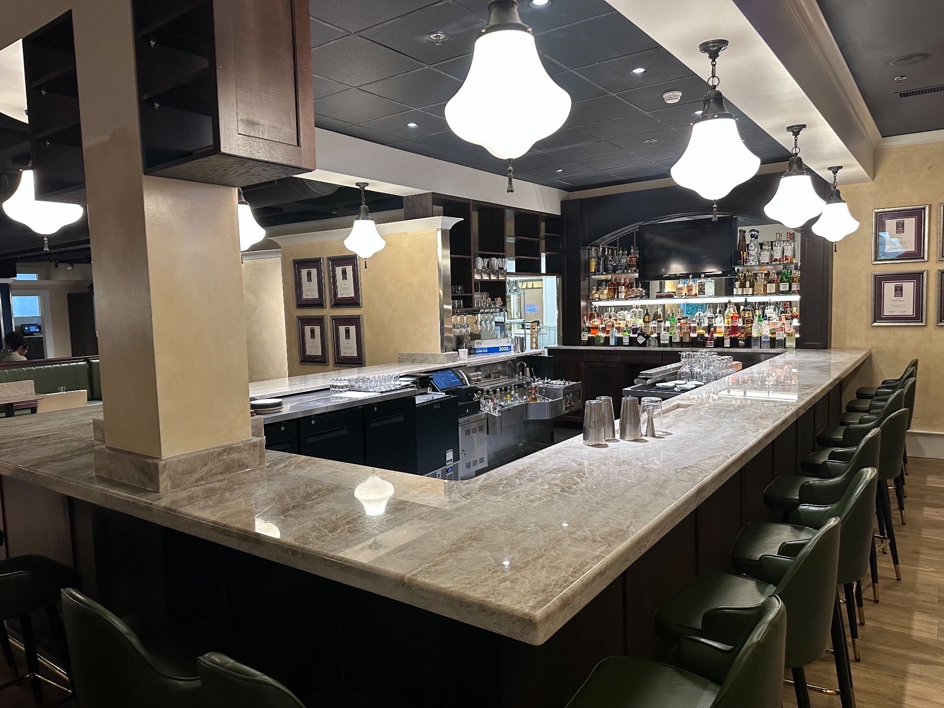 Bar with marble counter, dark wood, green barstools, overhead lights, and liquor bottles in the background.