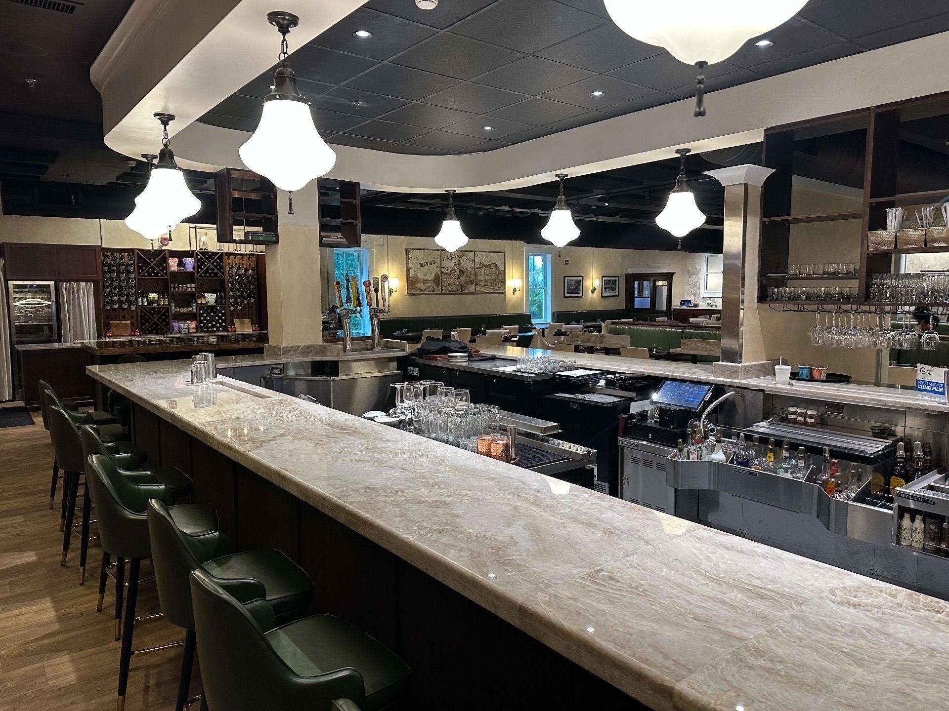 Empty restaurant bar with marble countertop, green bar stools, and overhead lamps.