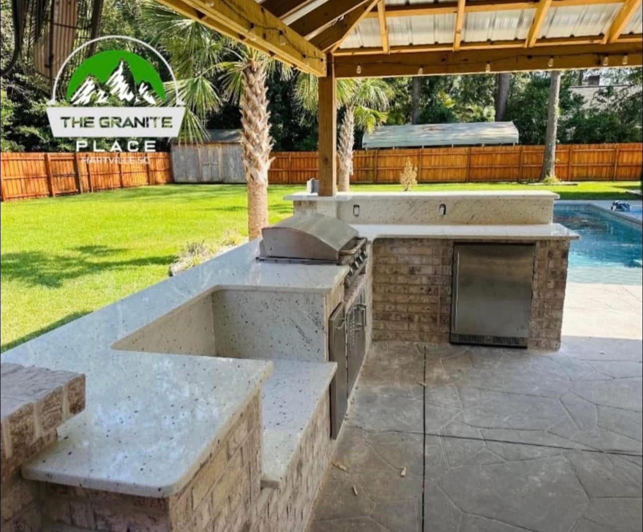 Outdoor kitchen with granite countertops, grill, refrigerator, and brick accents, beside a pool.