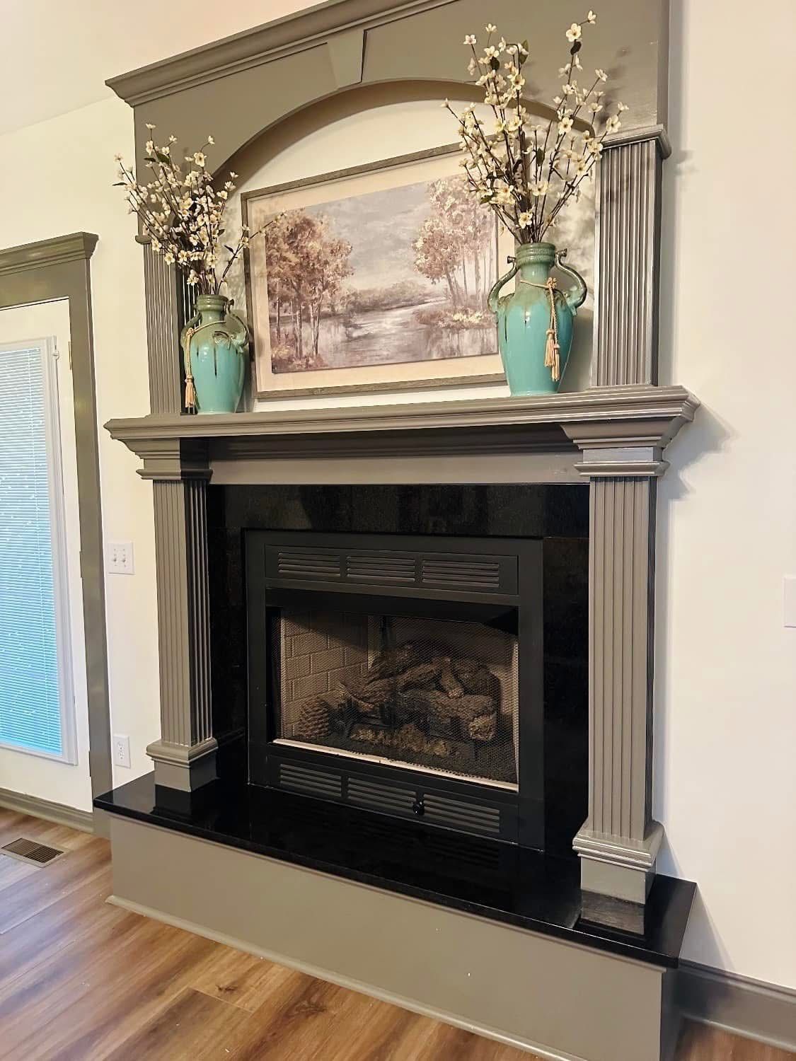 Fireplace with grey mantel, black surround, artwork, and turquoise vases with white flowers.