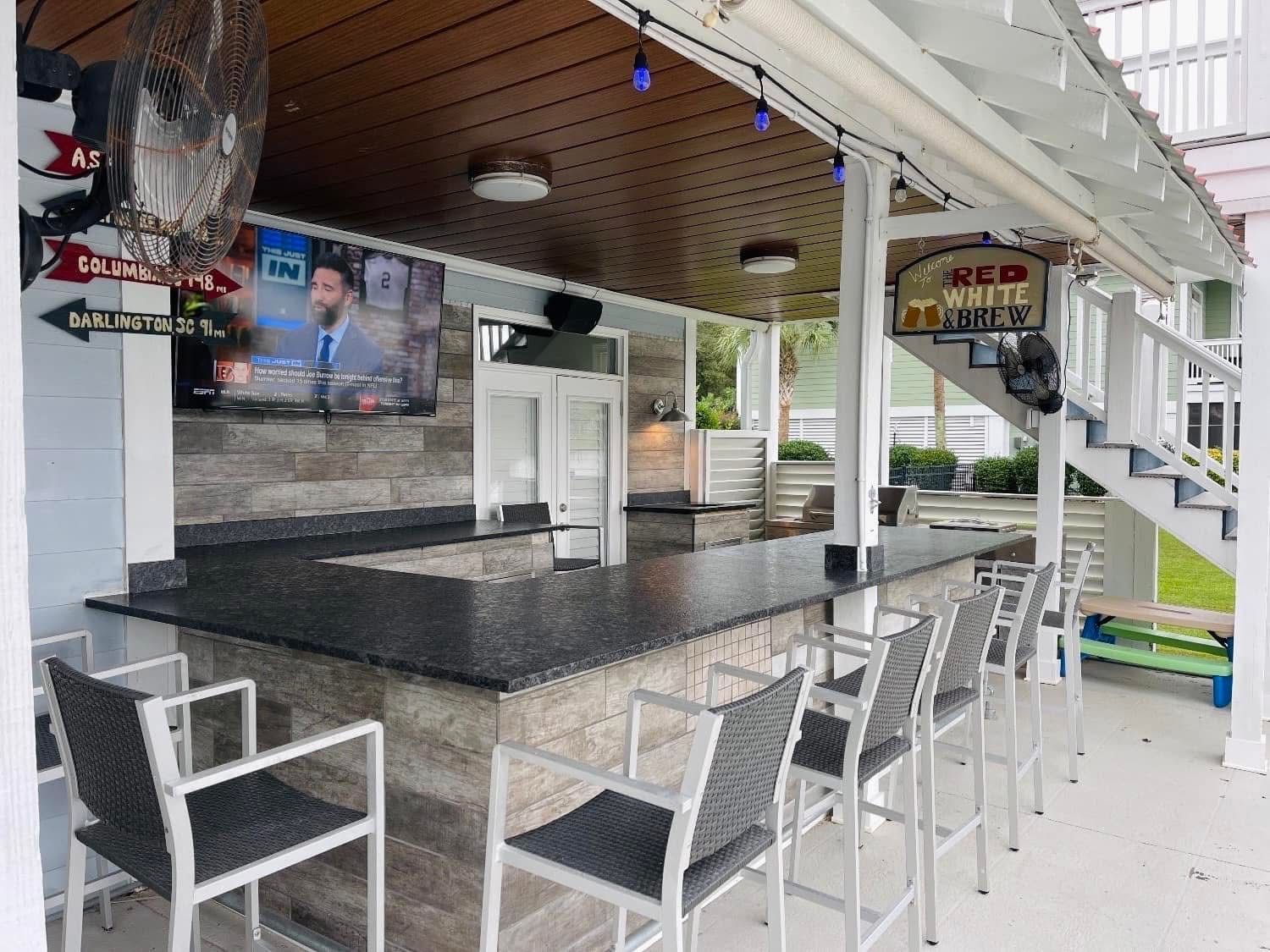 Outdoor bar with granite countertop, bar stools, TV, refrigerator, ceiling fan, and sign.