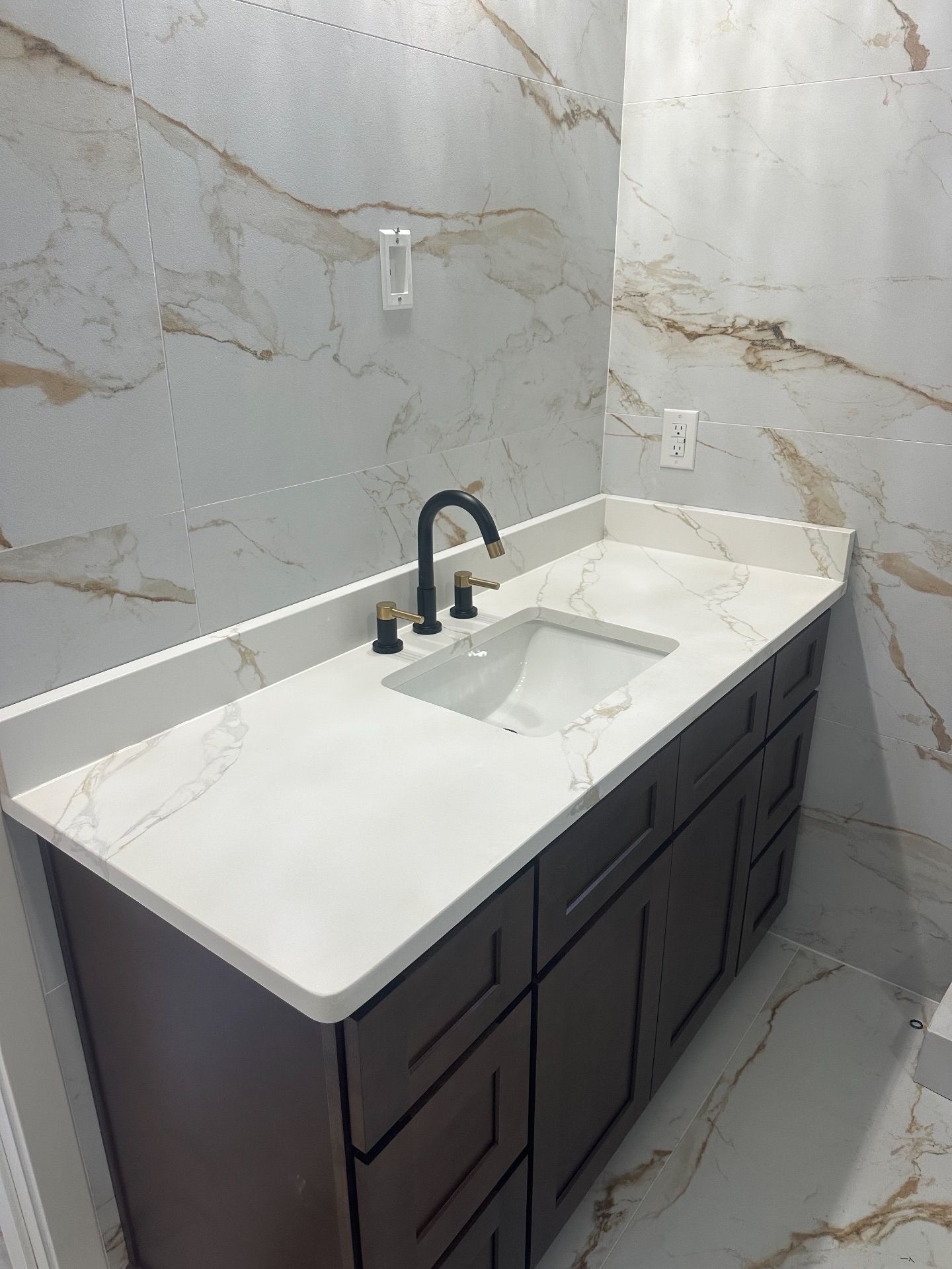 Bathroom vanity with dark brown cabinets, white countertop, and black faucet against marble-look tile walls and floor.