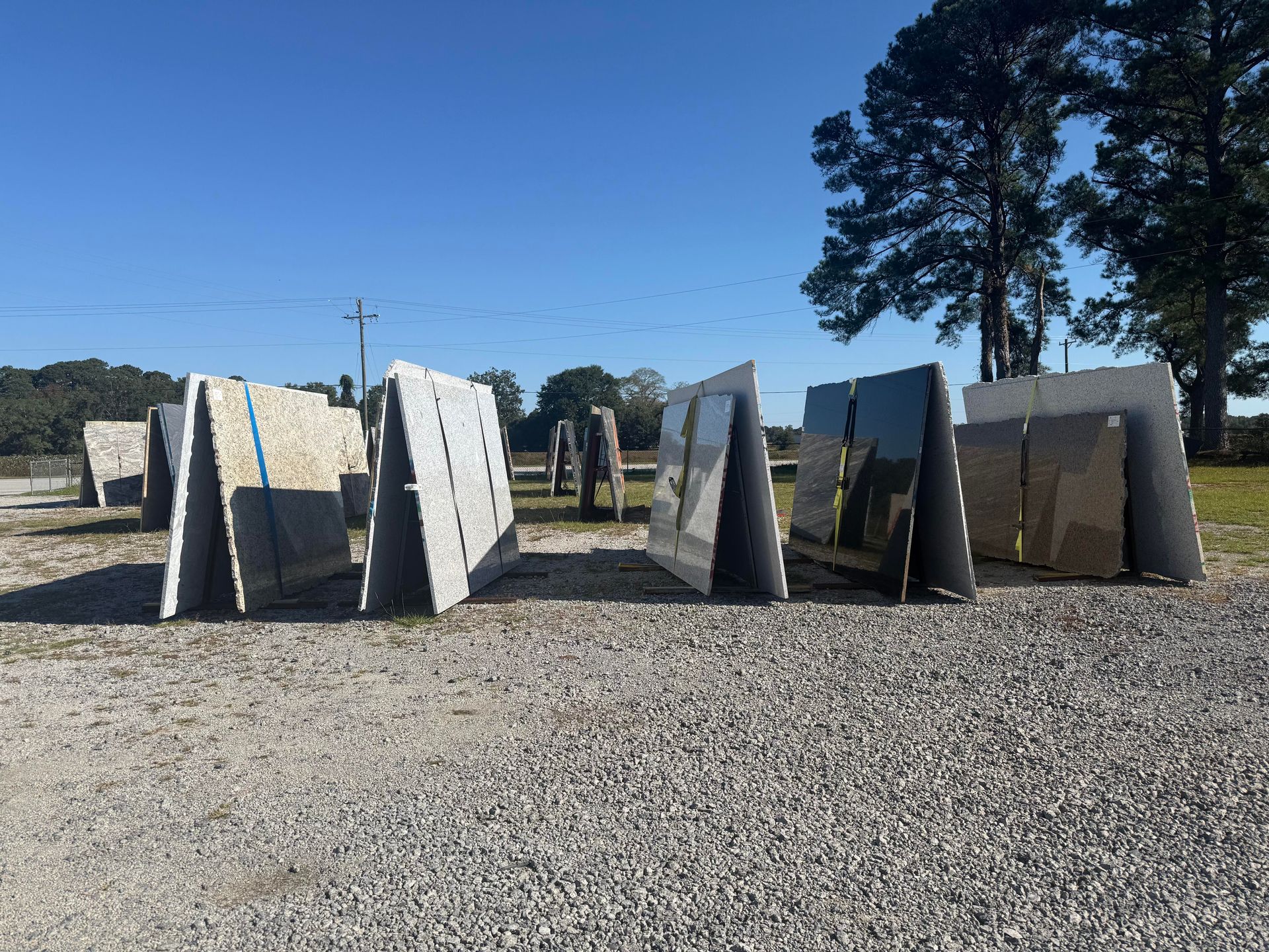 Slabs of stone displayed outdoors, leaning against A-frames, on a gravel lot under a blue sky.