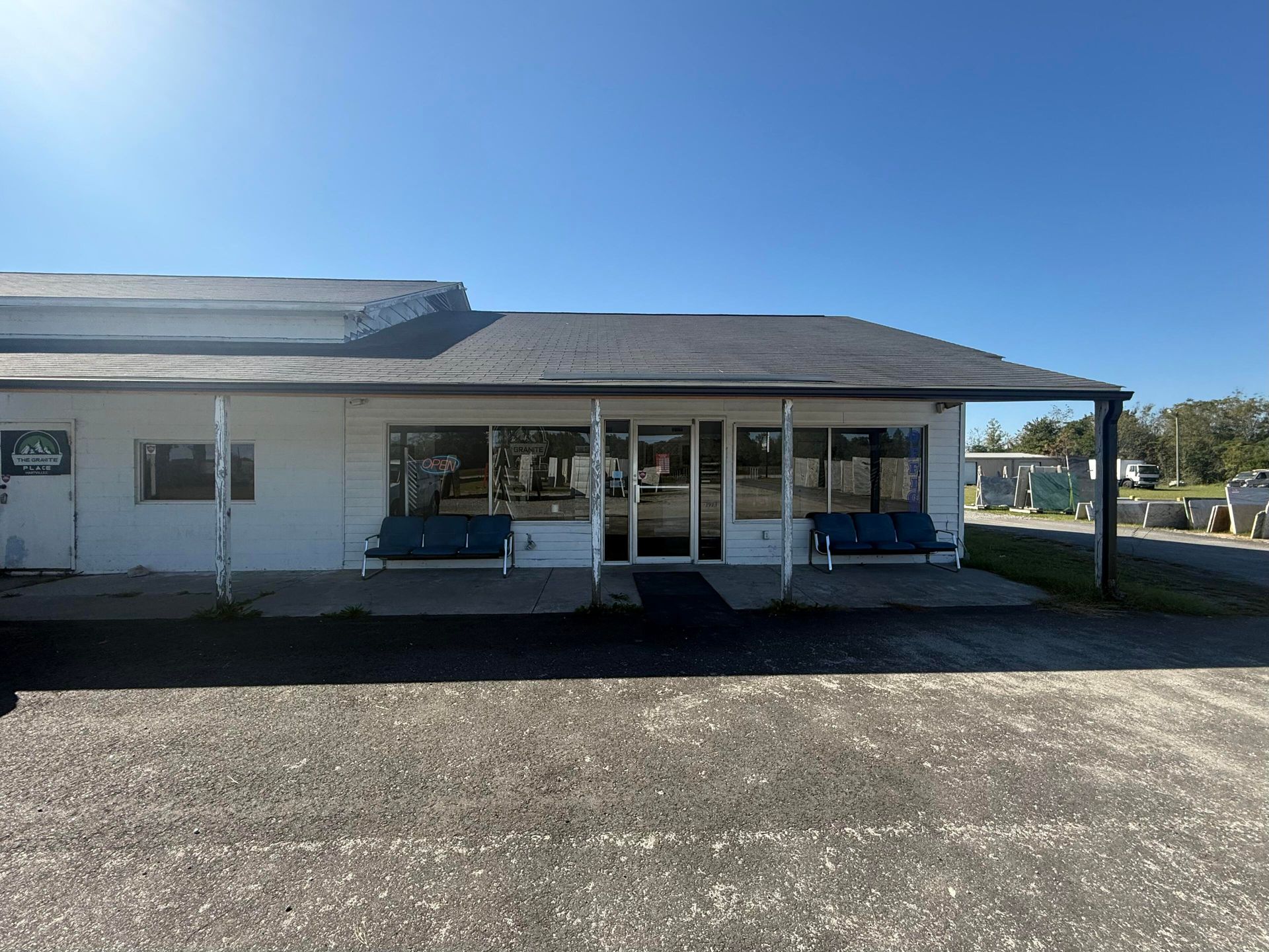 White building with dark roof. Glass doors, two blue benches, gravel lot, and clear blue sky.
