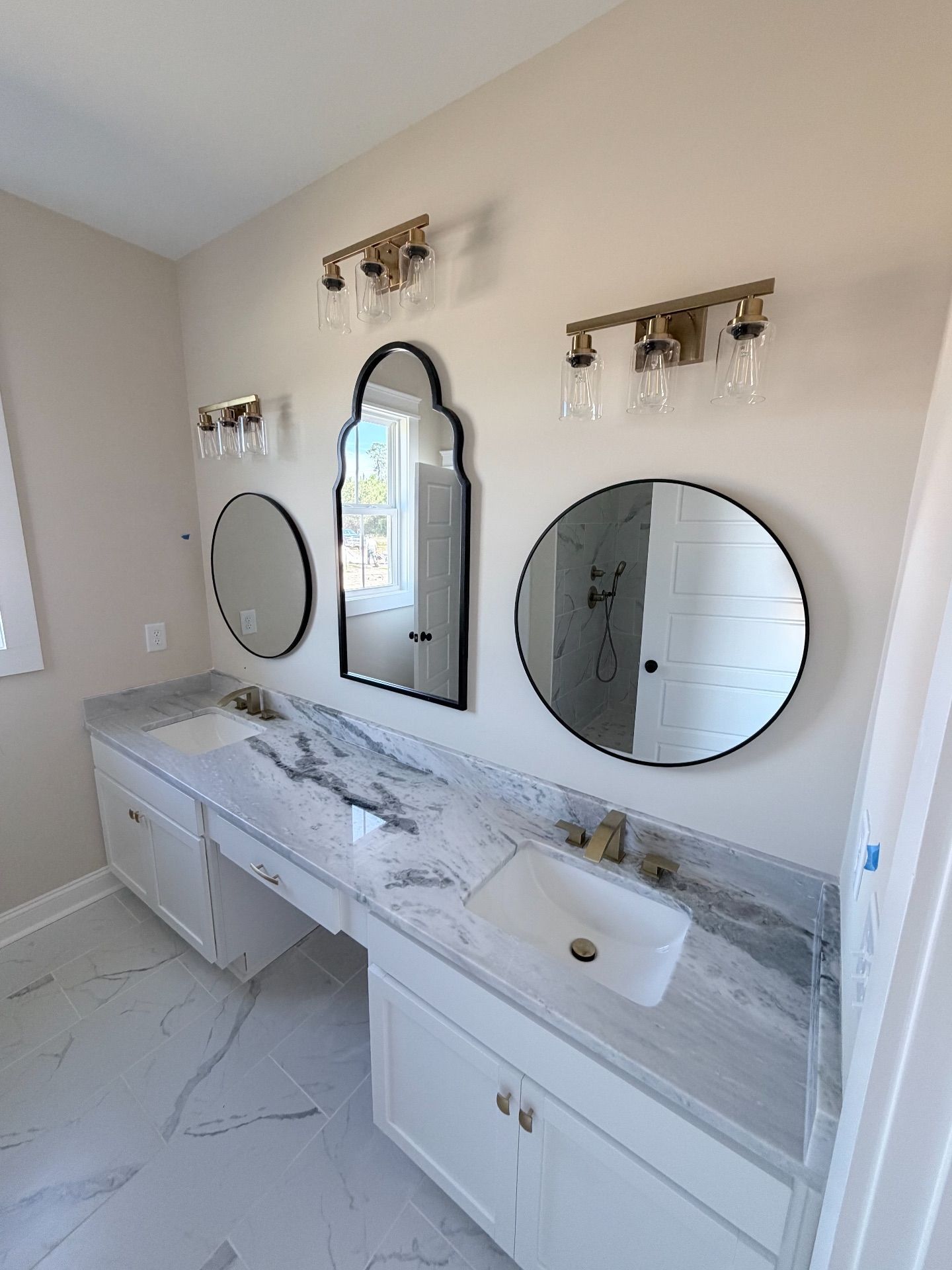 Bathroom with white cabinetry, marble countertops, and three mirrors with gold sconces.