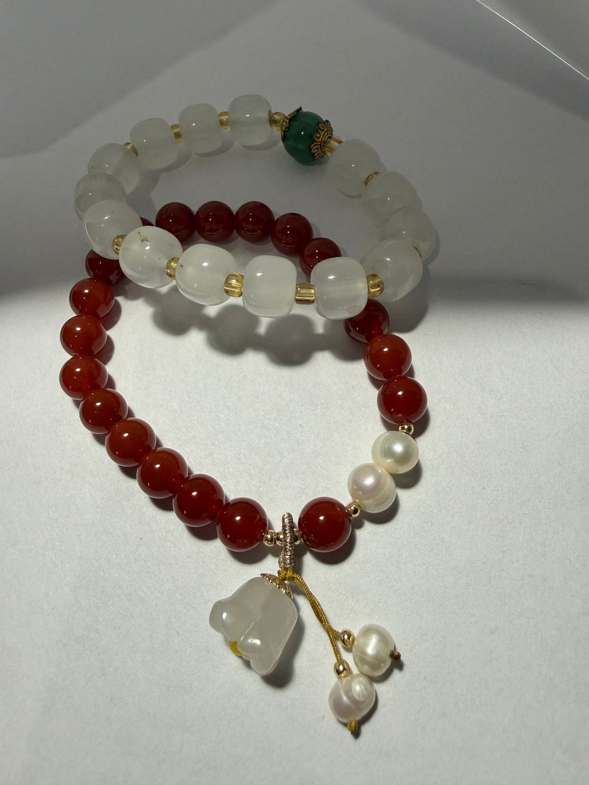 Dos pulseras de cuentas: una marrón rojiza y otra blanca. Detalles dorados y un colgante de flor con borlas de perla.