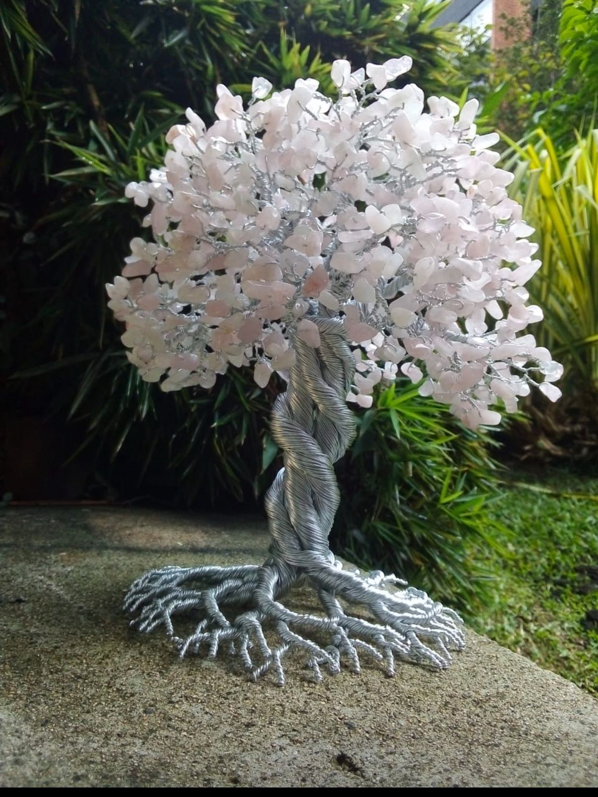 Escultura de árbol de alambre de plata con hojas de cristal rosa, sobre una superficie texturizada, con vegetación de fondo.