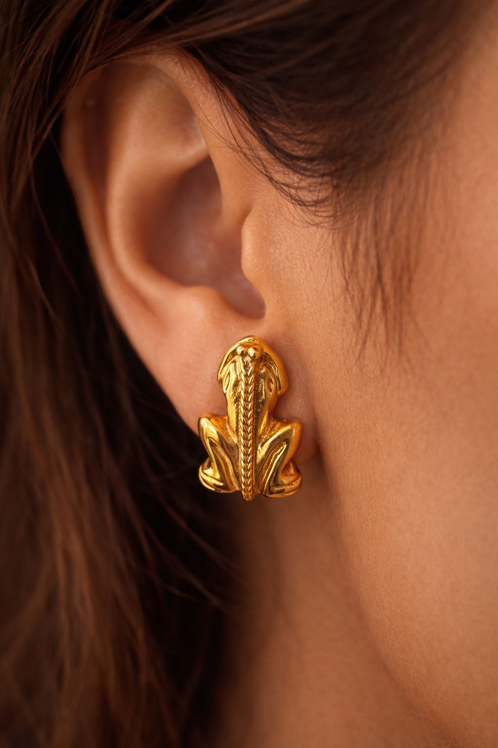 Una persona lleva en el lóbulo de la oreja un pendiente texturizado de color dorado con forma de rana sentada.