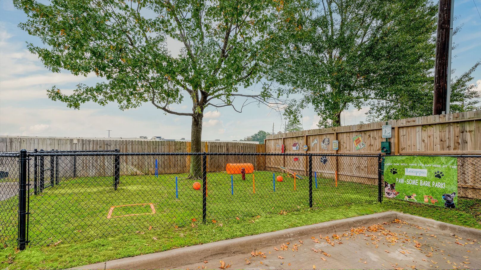 A fenced-in dog park with agility equipment, grassy ground, and trees on a sunny day at Lexington at Champions in Houston, TX..