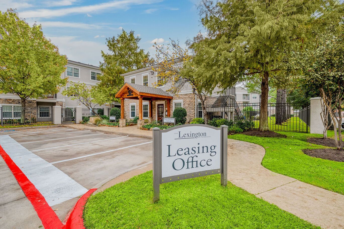A Leasing Office sign stands in front of a tan apartment building with a wooden entrance canopy and surrounding trees at Lexington at Champions in Houston, TX..