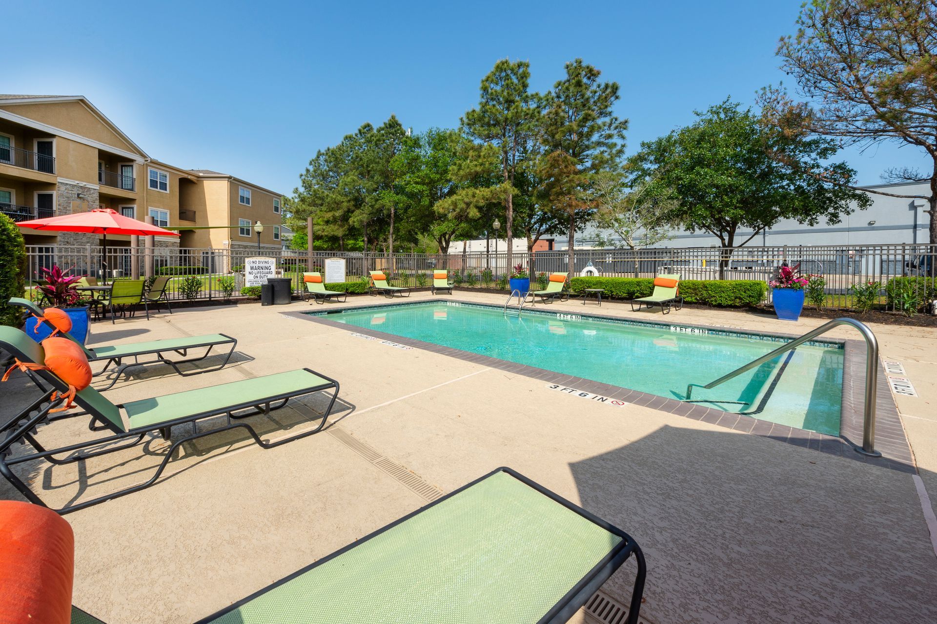 Outdoor swimming pool with lounge chairs and green trees under a bright blue sky at an apartment complex at Lexington at Champions in Houston, TX..