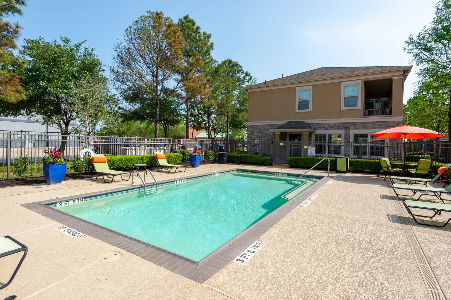 A rectangular swimming pool surrounded by lounge chairs, an orange umbrella, and landscaping with a building in the back at Lexington at Champions in Houston, TX..