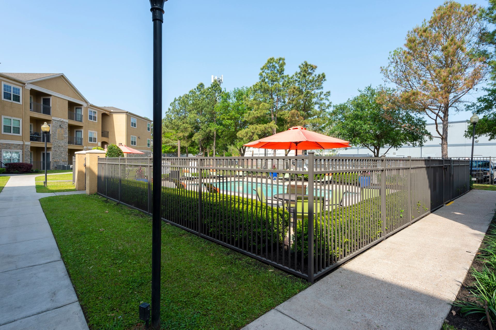 An apartment complex pool area with a metal fence, green lawn, sidewalk, and an orange umbrella on a sunny day at Lexington at Champions in Houston, TX..