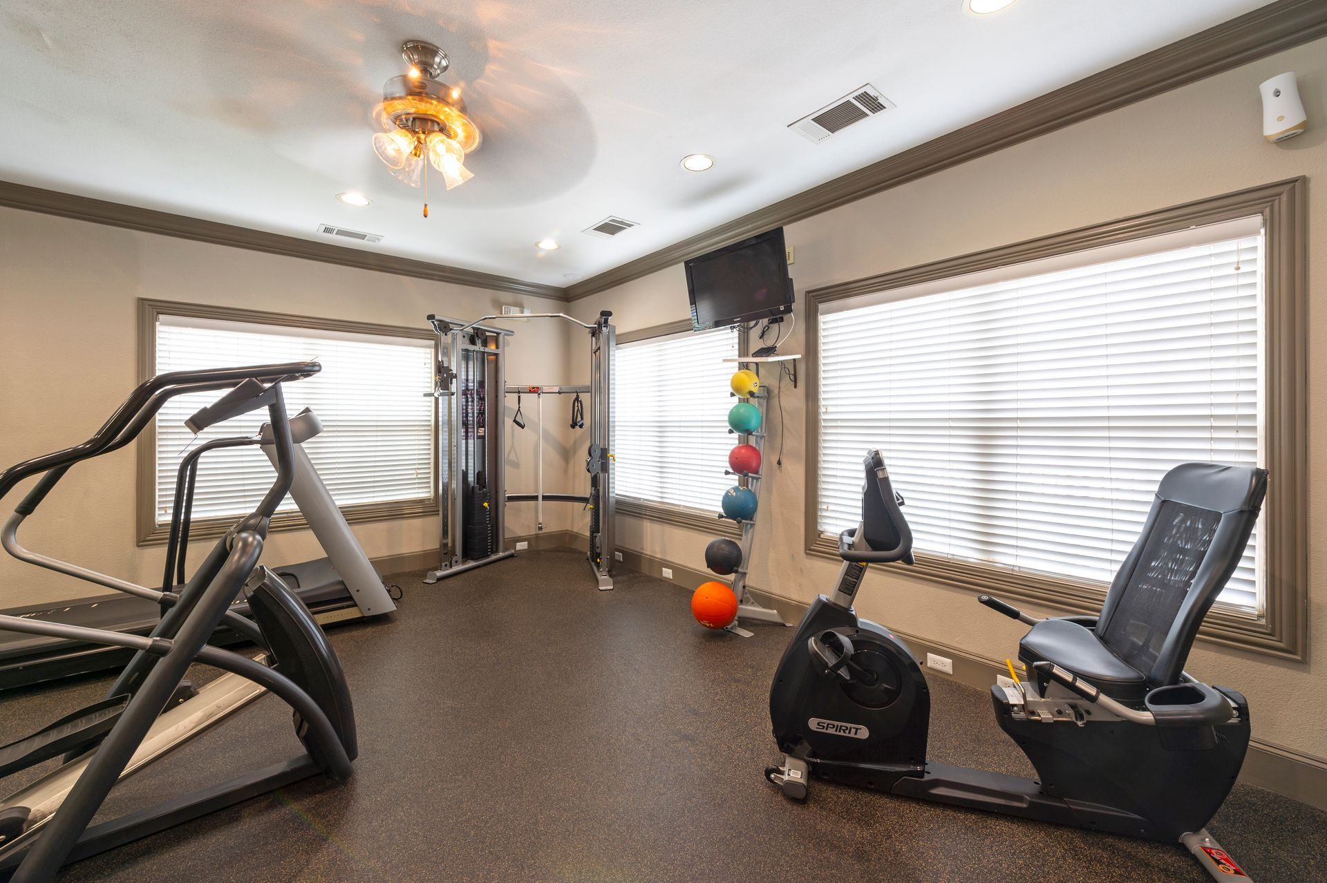 A residential fitness room with a stationary bike, elliptical, and a vertical rack of exercise balls near window blinds at Lexington at Champions in Houston, TX..