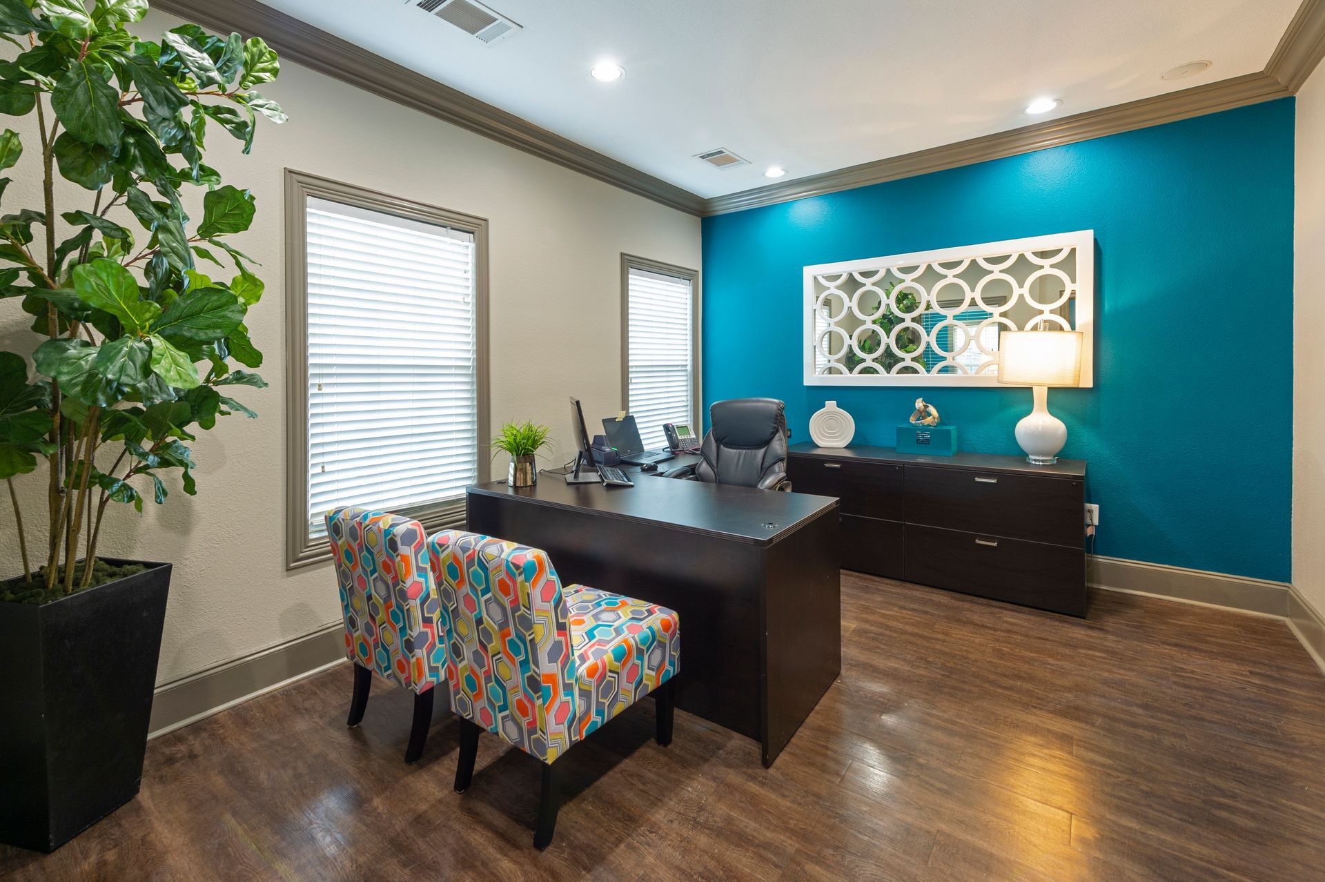 A home office featuring a dark wood desk, two colorful patterned chairs, a tall indoor plant, and a bright teal wall at Lexington at Champions in Houston, TX..