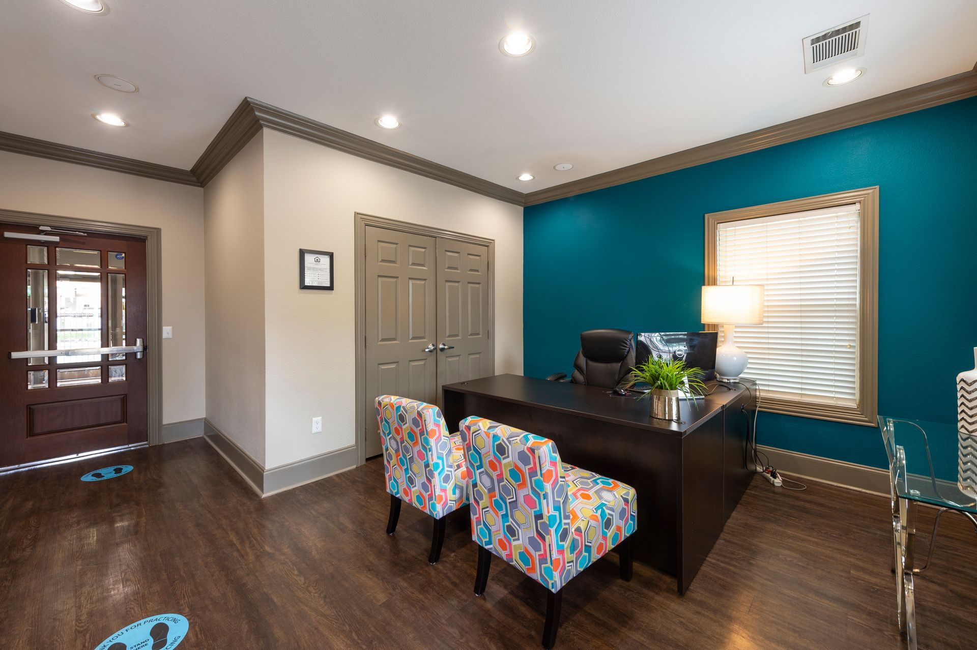 An office interior with a dark wood desk, two patterned armchairs, a turquoise wall, and a wooden entry door at Lexington at Champions in Houston, TX..