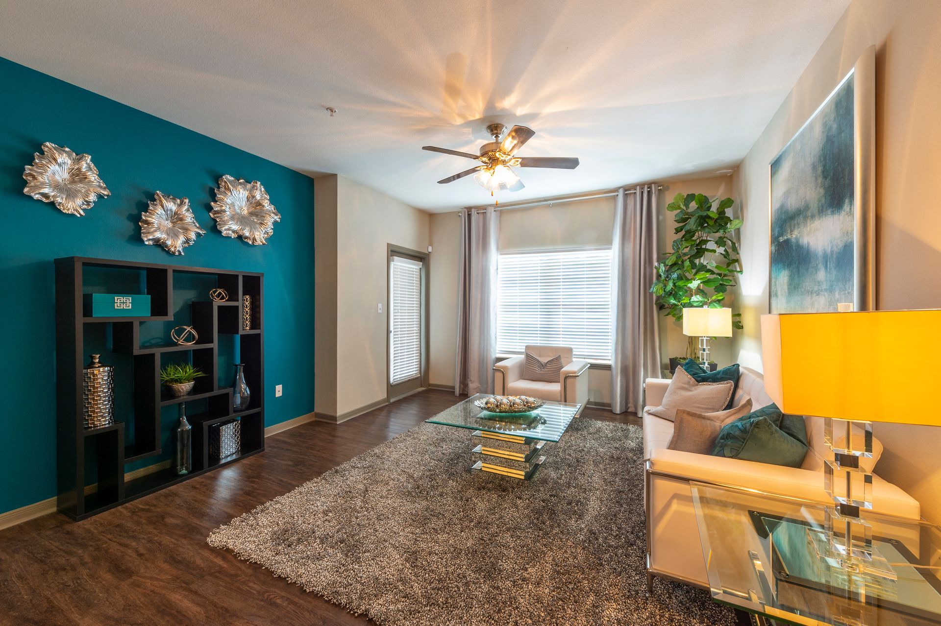 Modern living room with a teal wall, a dark bookshelf, a large rug, a sofa, and a yellow lamp at Lexington at Champions in Houston, TX..