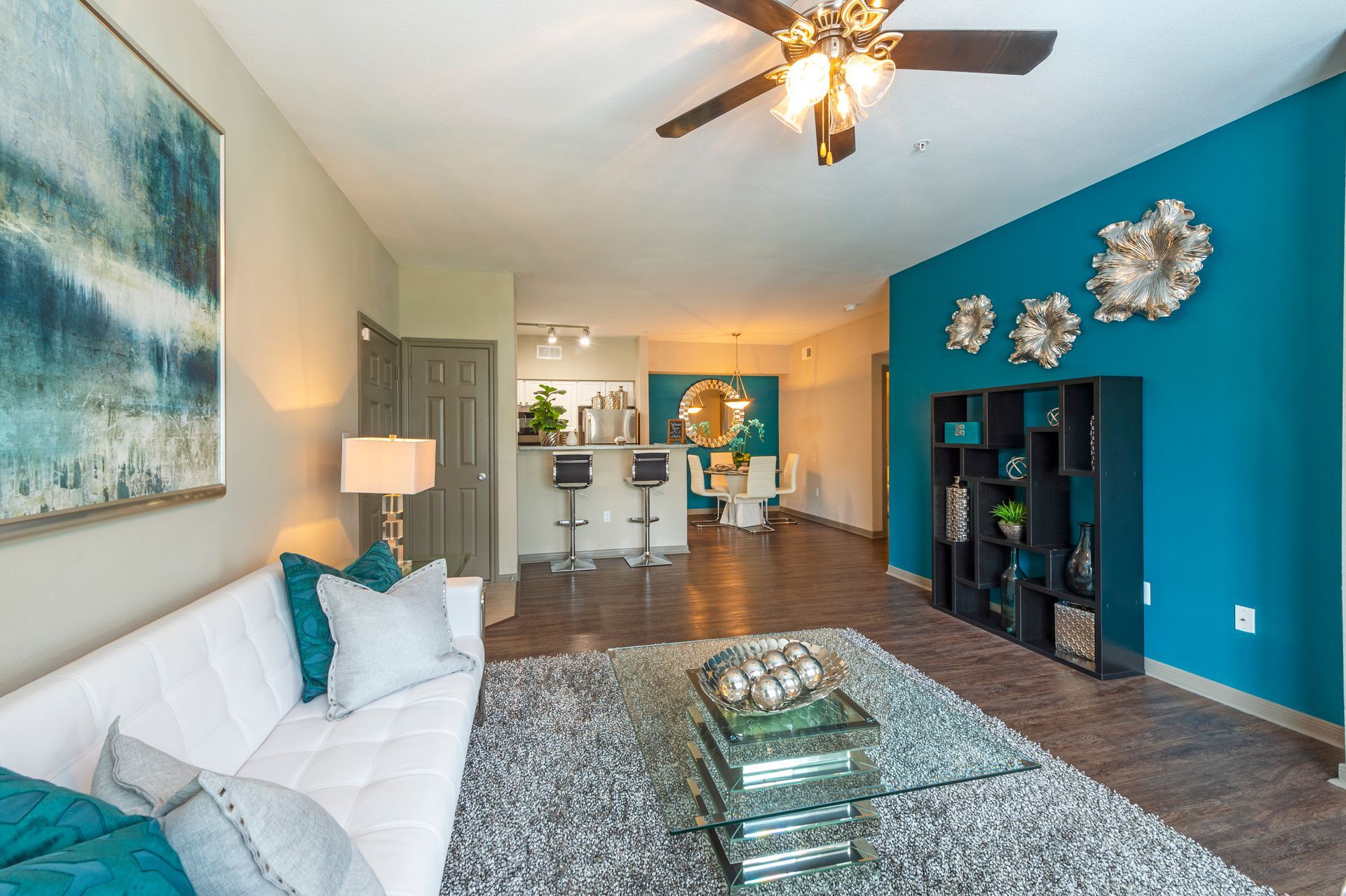 A living room with a white sofa, teal accent wall with silver decor, grey rug, glass coffee table, and open-plan kitchen at Lexington at Champions in Houston, TX..