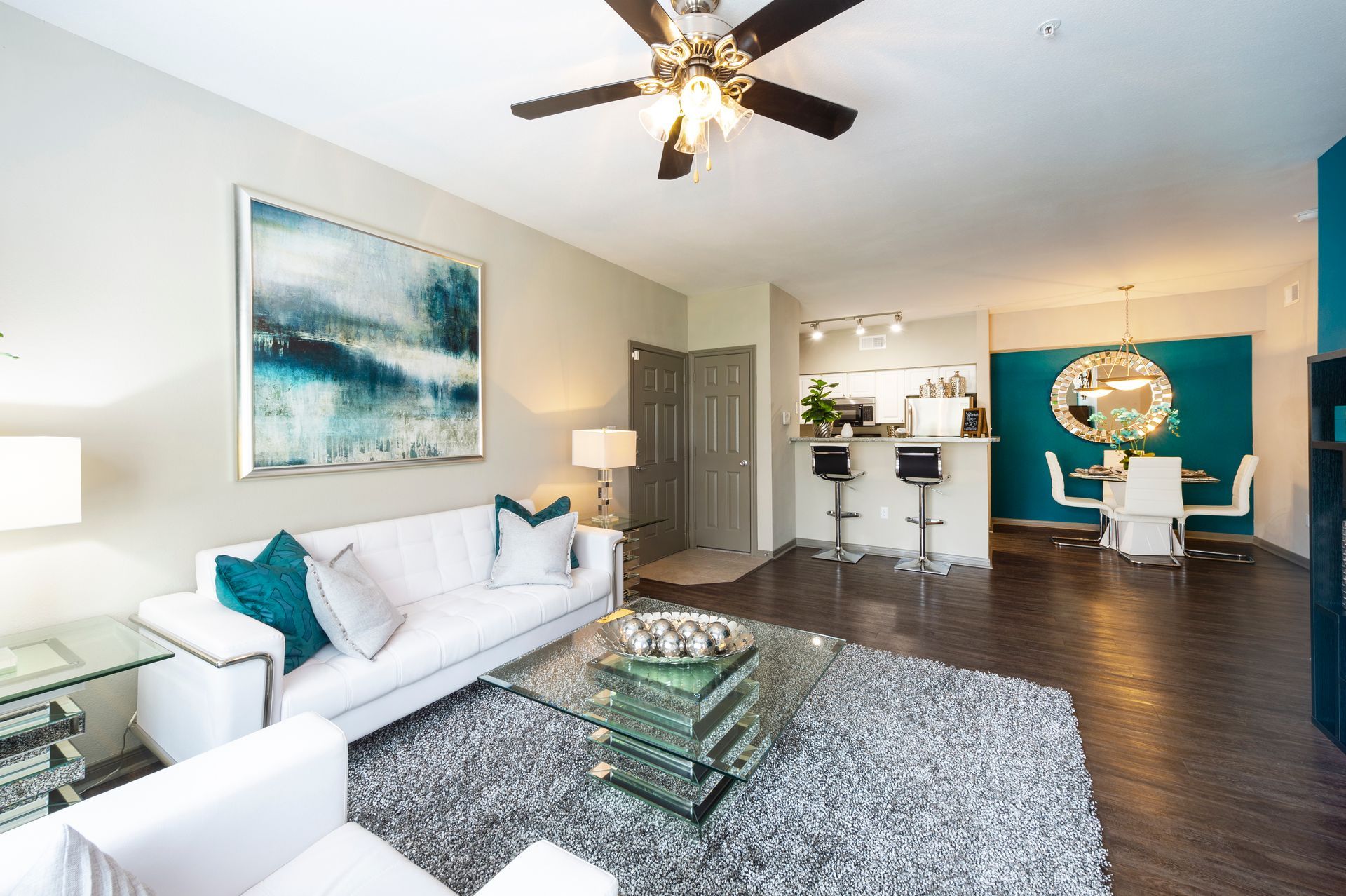 A modern living room with a white sofa, glass coffee table, grey rug, and an open kitchen with bar stools in the back at Lexington at Champions in Houston, TX..