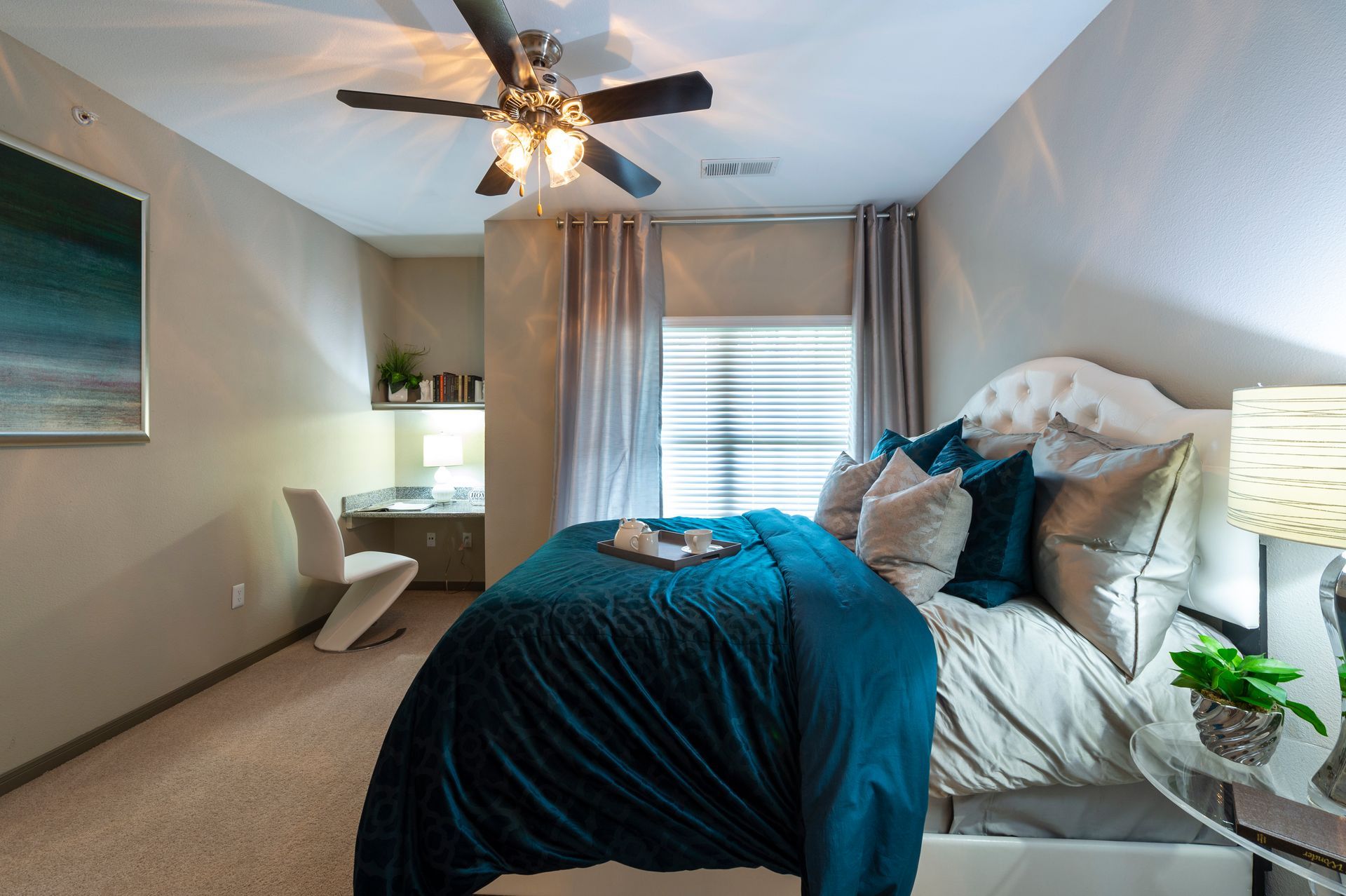 A modern bedroom featuring a bed with deep teal bedding, beige walls, a desk area, and a ceiling fan at Lexington at Champions in Houston, TX..
