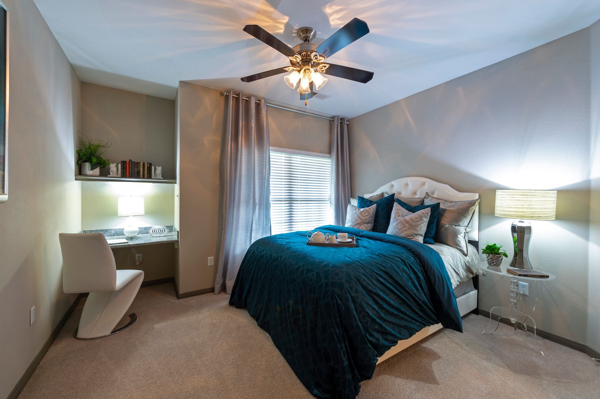 A cozy bedroom featuring a teal comforter on a bed, a ceiling fan, a window with curtains, and a small desk with a chair at Lexington at Champions in Houston, TX..