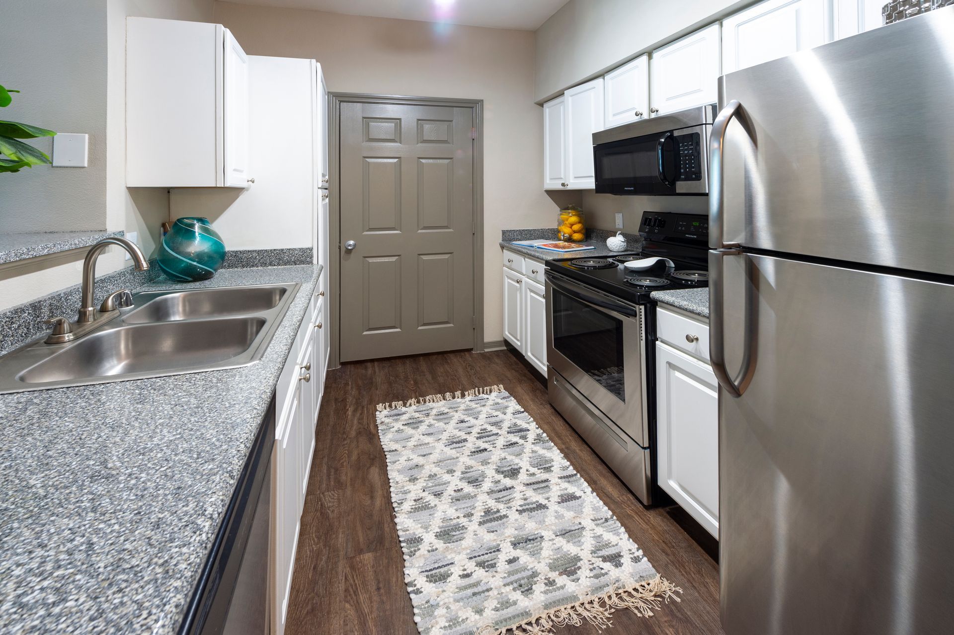A kitchen with white cabinets, gray granite countertops, stainless steel appliances, and a patterned rug on wood floors at Lexington at Champions in Houston, TX..