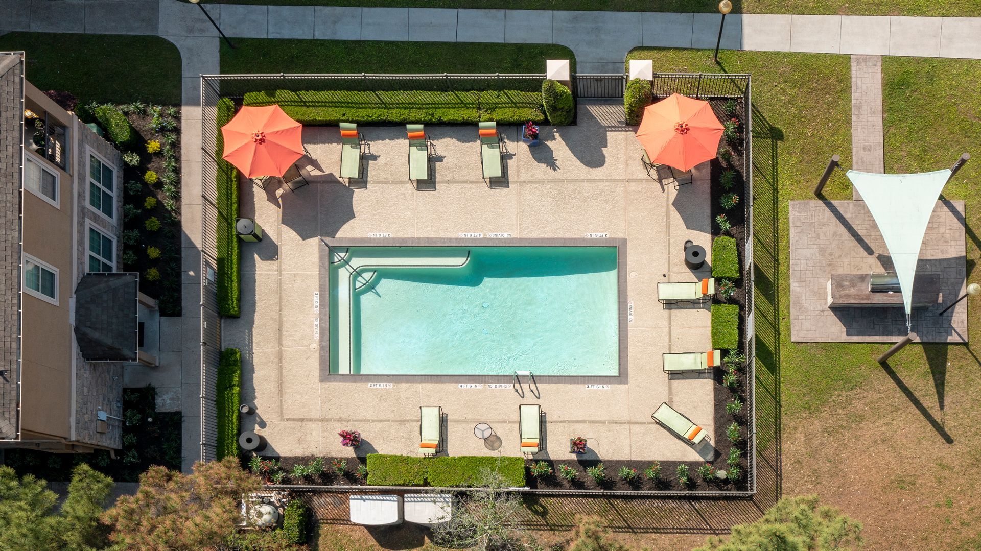 Overhead view of a rectangular pool with lounge chairs, orange umbrellas, and a shaded picnic area.