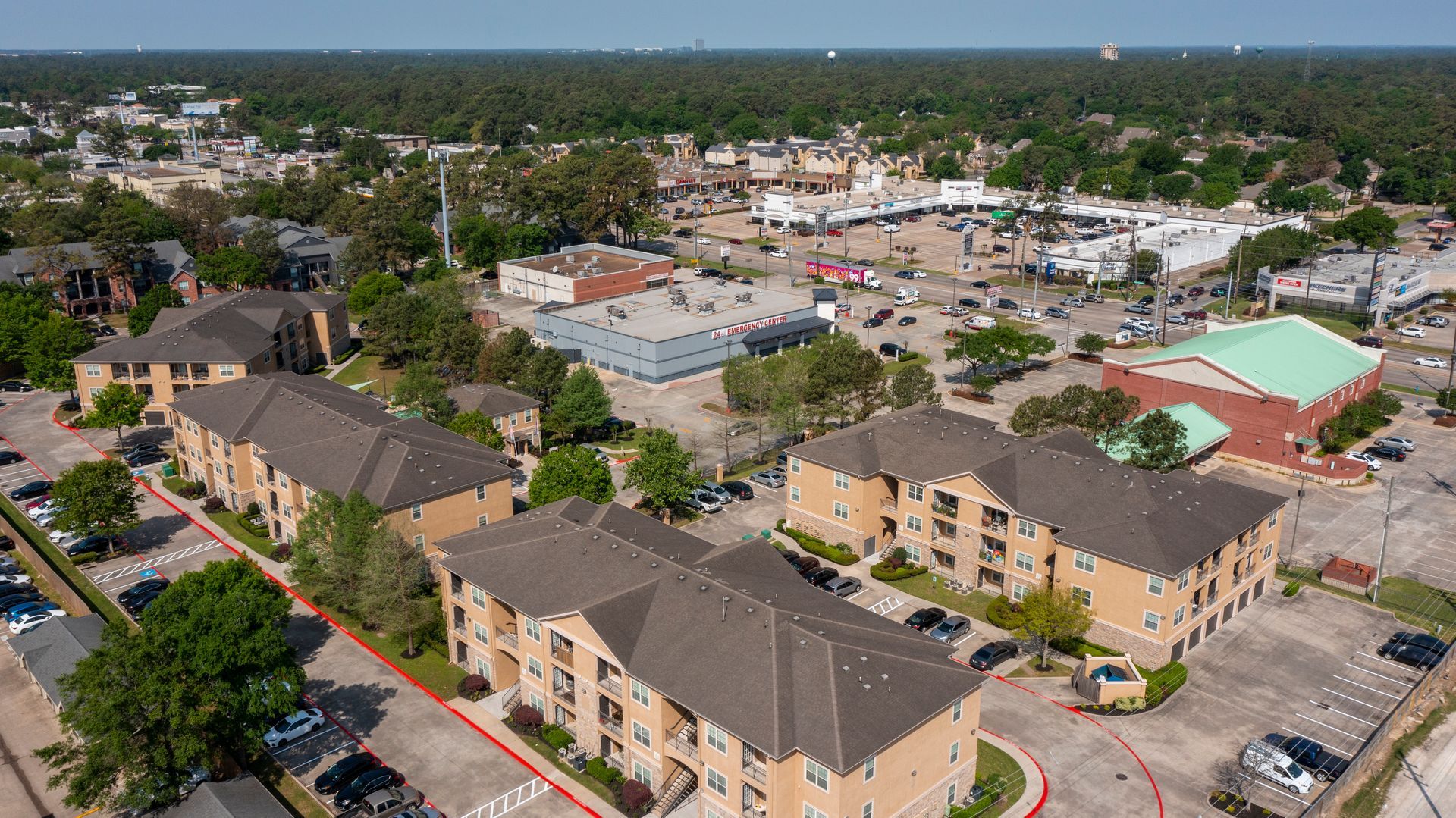 Aerial view of a residential apartment complex with brown roofs and a nearby shopping center surrounded by green trees at Lexington at Champions in Houston, TX..