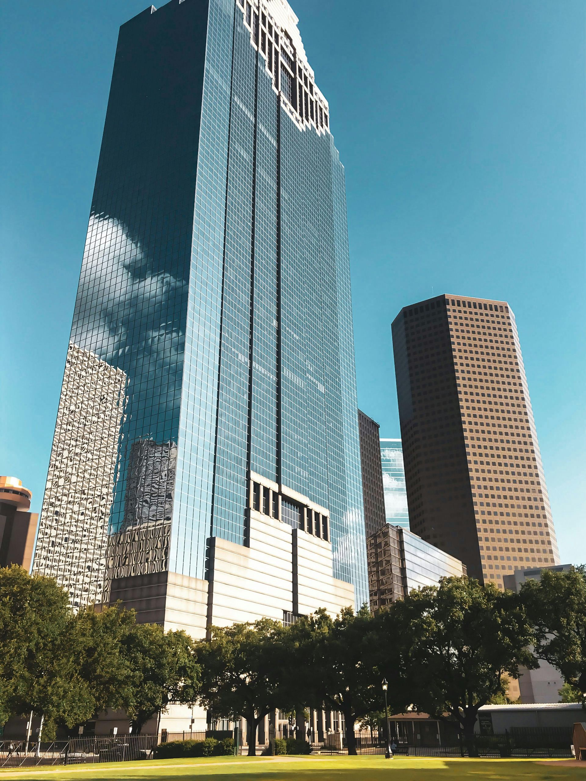 A tall, modern glass skyscraper reflects a cloudy sky next to a tan stone building, all viewed from a park with trees at Lexington at Champions in Houston, TX..